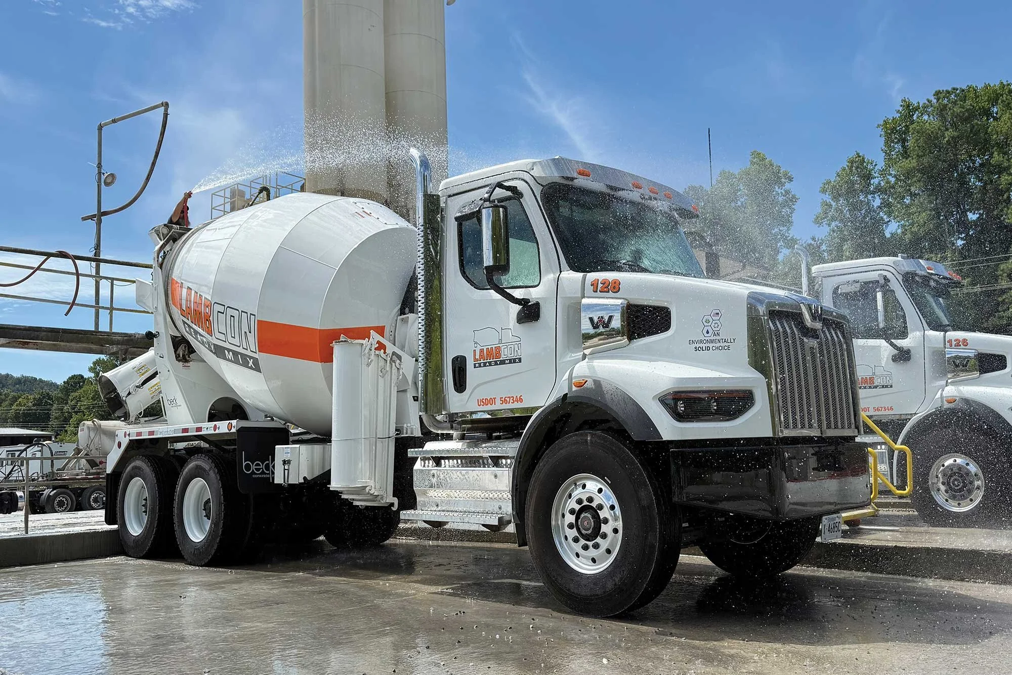 A white cement mixer truck with orange and black markings and the word 'LAMBEcon' on the drum, water spray from a hose, and a worker on a platform, against a blue sky and green trees.