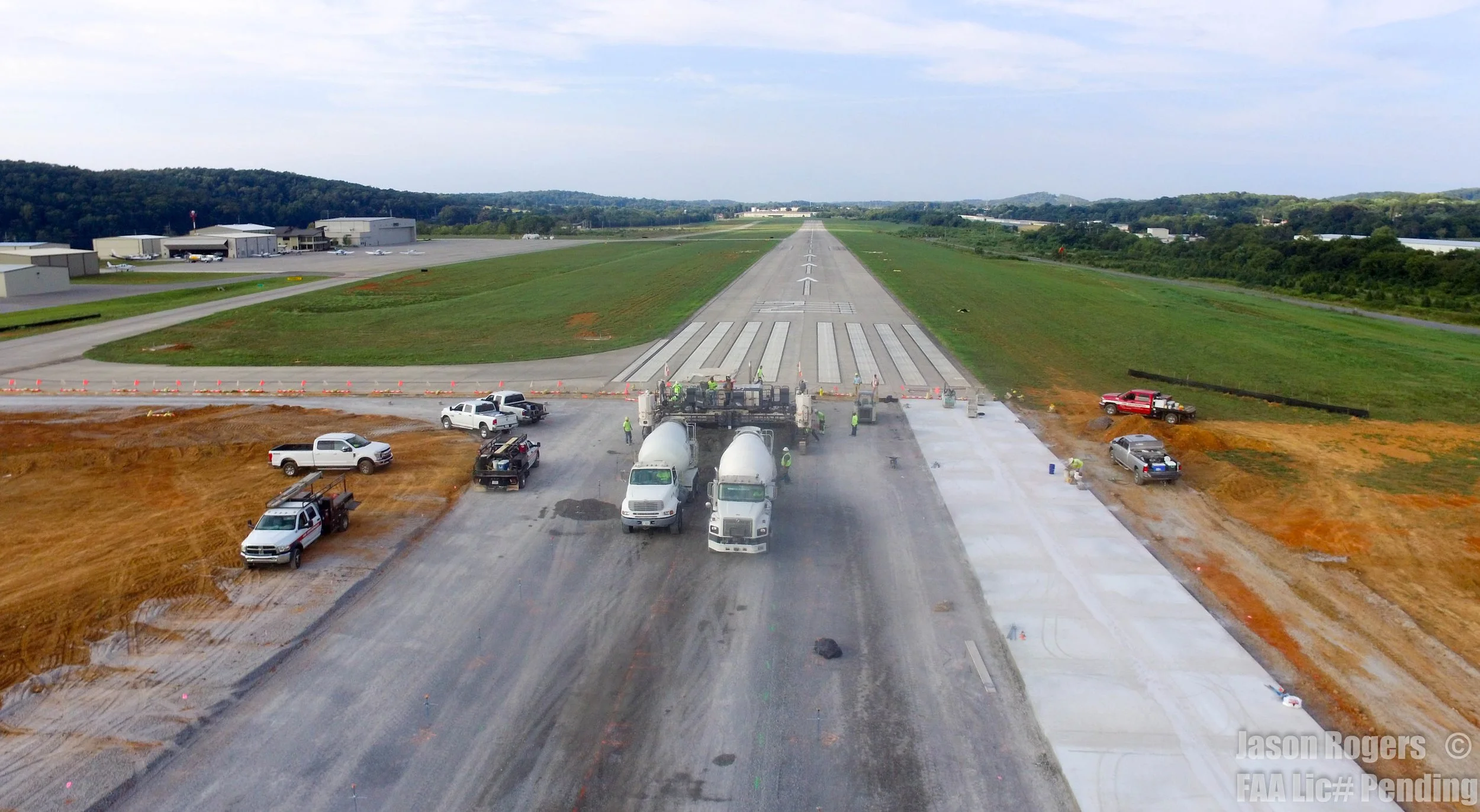 Construction workers and vehicles are paving a new runway at an airport, with trees and airport buildings in the background.