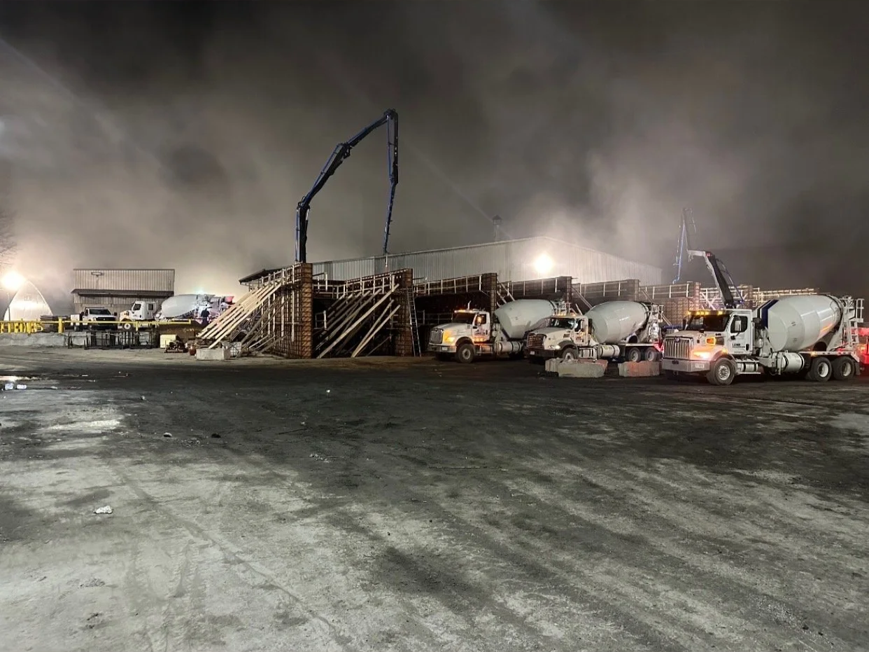 Construction site at night with concrete mixer trucks, scaffolding, and cranes, surrounded by dirt and smoke.