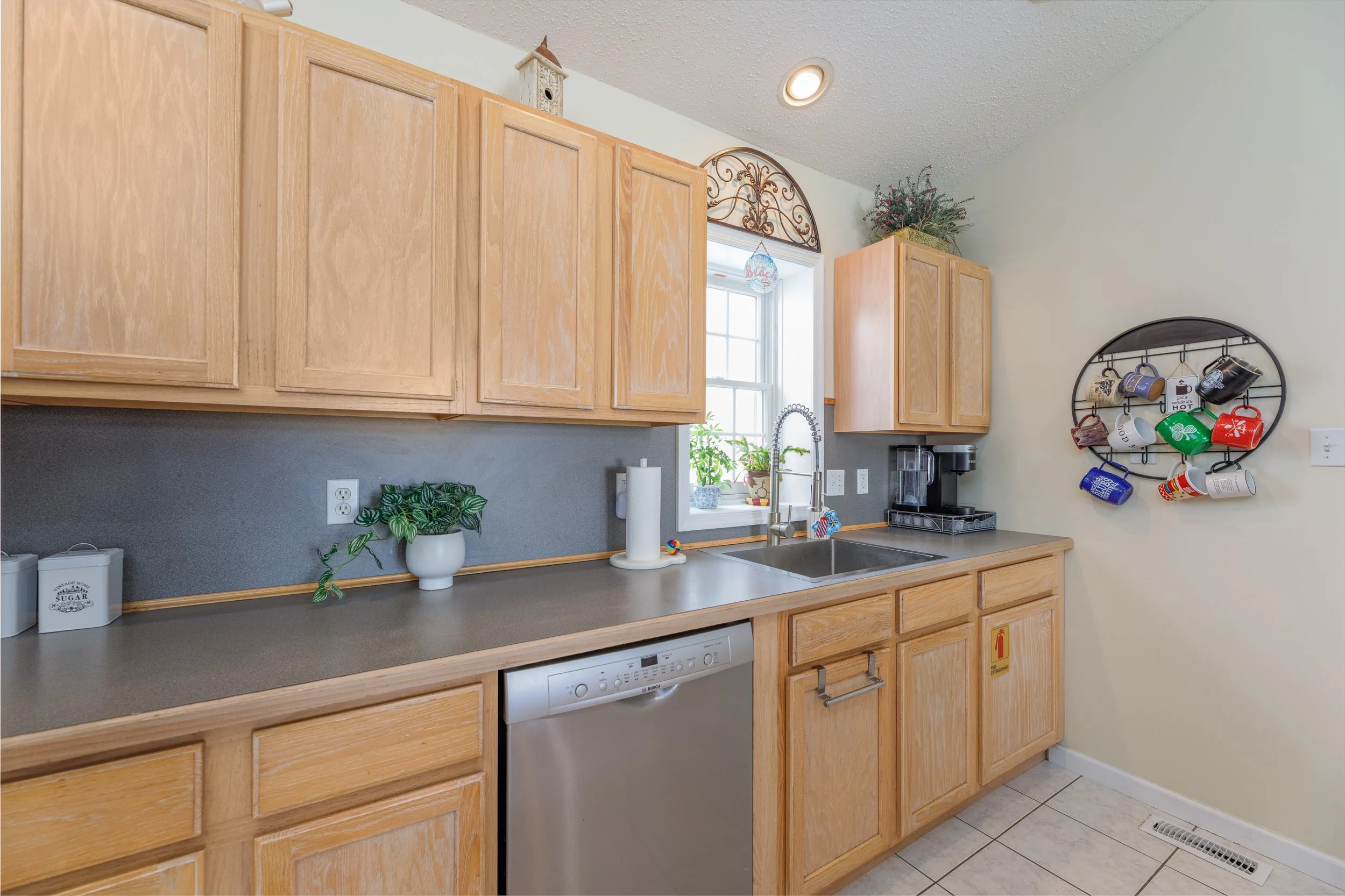 Kitchen with light wooden cabinets, gray countertop, window with plants, hanging mug holder with colorful mugs, dishwasher, and a small decorative birdhouse and plant above the cabinets.