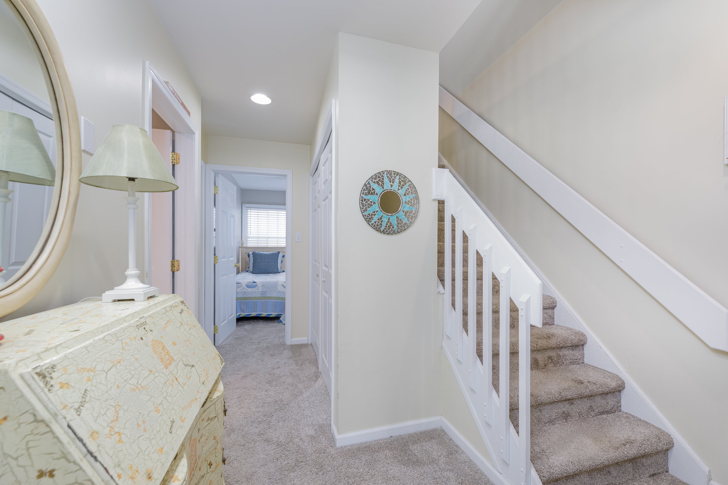View of a hallway with carpeted stairs on the right, a white wall with a round decorative mirror, a doorway leading to a bedroom with a bed and window, a white bookshelf, a corner with a lamp and a mirror, and a patterned chest or trunk in the foregr