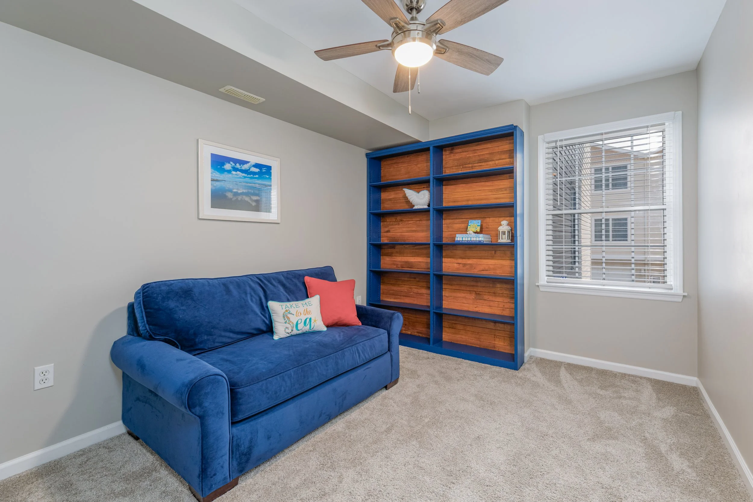 A living room with a blue velvet couch, a throw pillow with the words "Take me to the sea", a wooden bookshelf painted blue, a framed photo of a blue sky and clouds, a window with white blinds, a ceiling fan, and beige carpet.