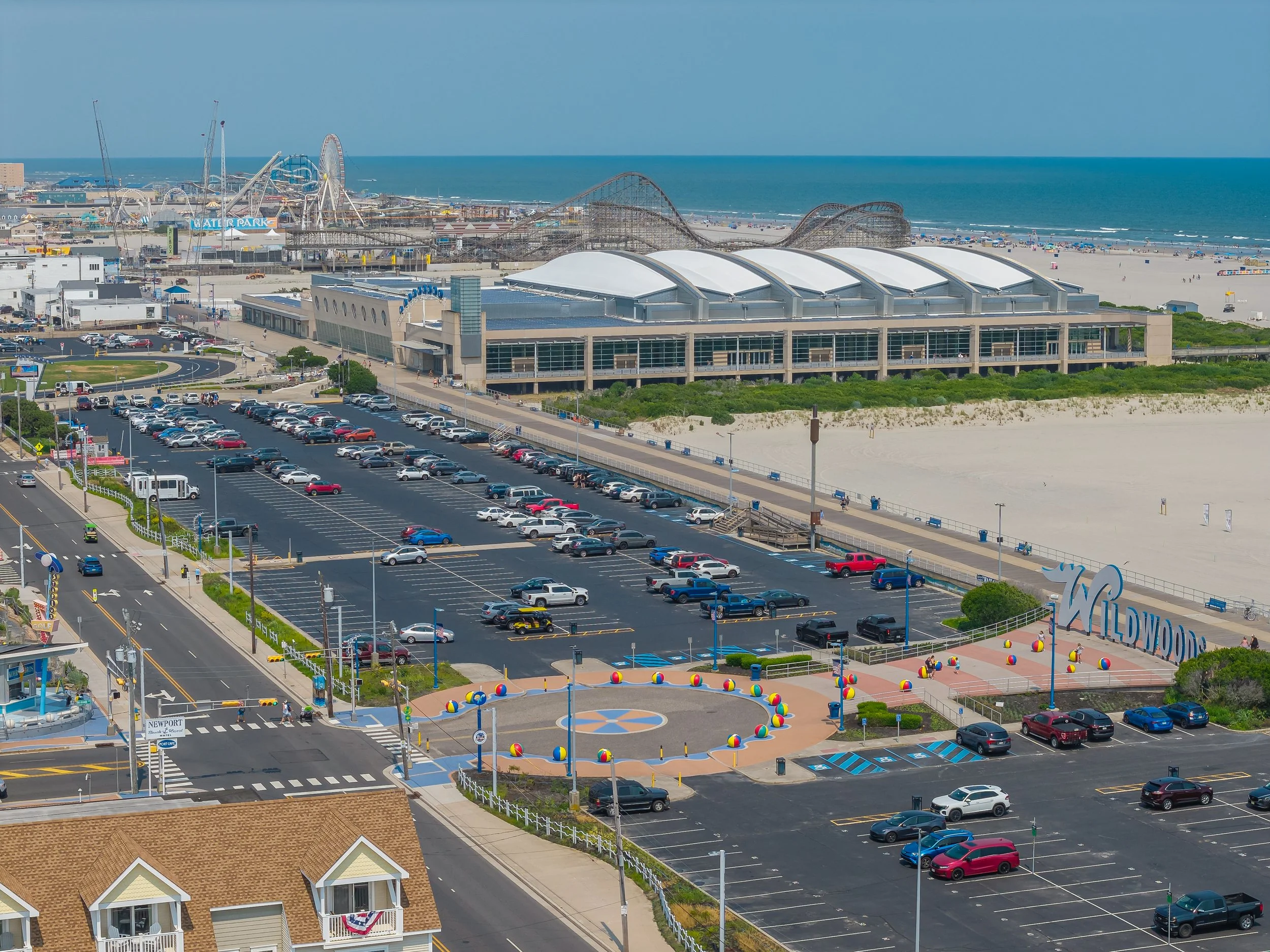 Aerial view of an amusement park and beach with roller coasters, parking lot, and colorful beach umbrellas along the shoreline.