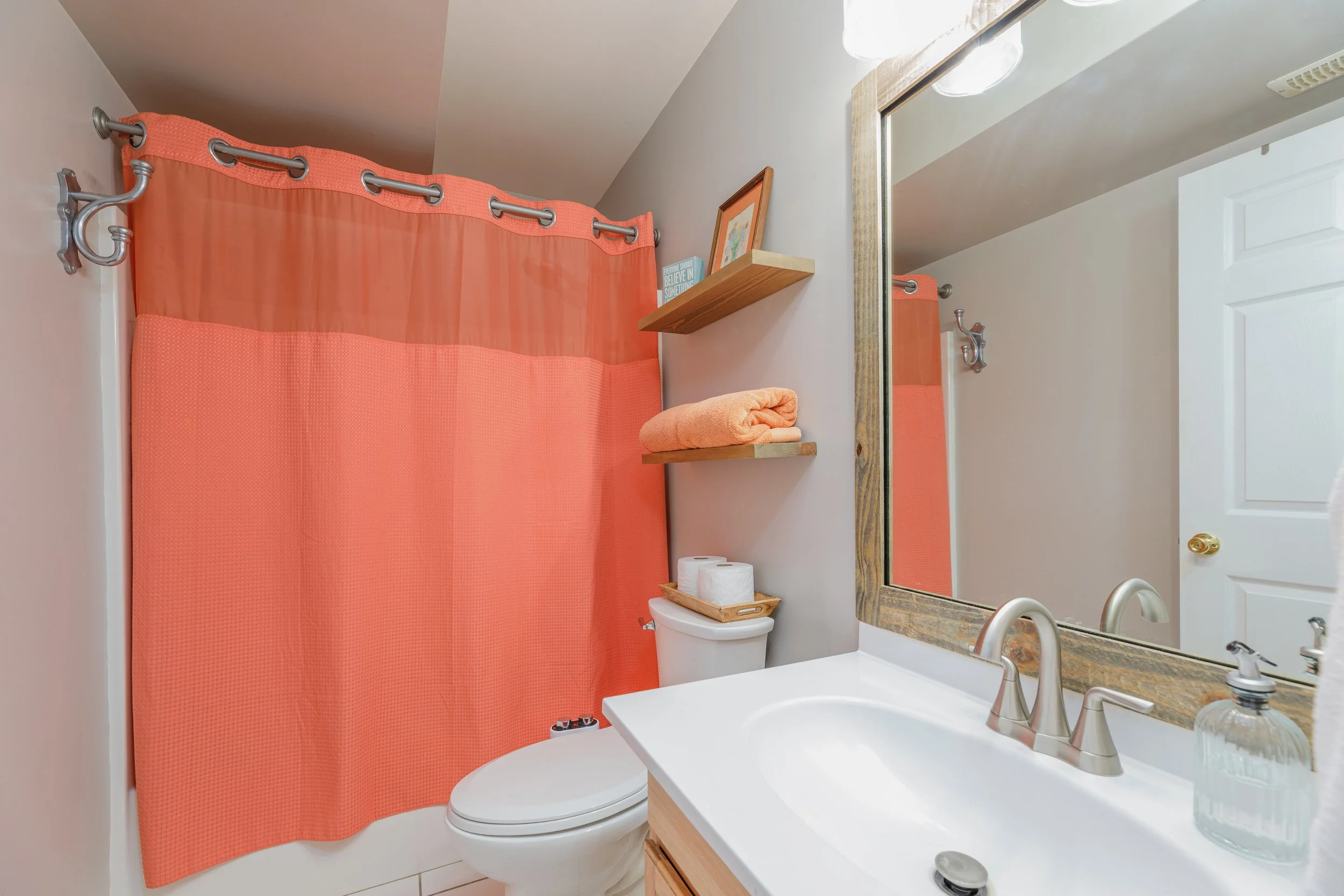 A bathroom with white walls and a white toilet, a mirror with a wooden frame, a white sink with a silver faucet, two shelves with a framed picture and an inspirational book, a coral-colored shower curtain, a light-colored towel, and some toiletries.
