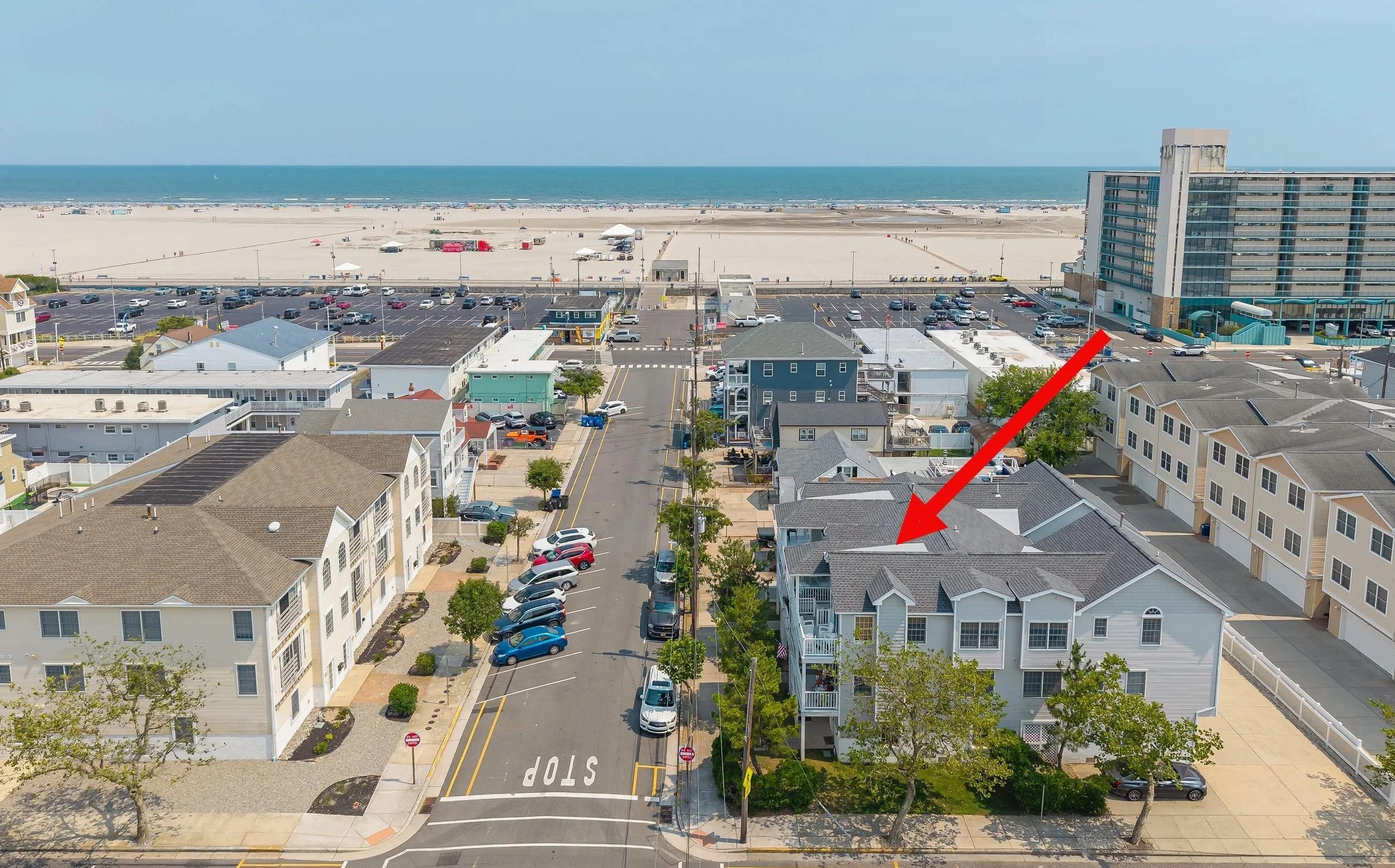 Aerial view of a beach town showing residential houses, a parking lot, and the beach with the ocean in the background. An arrow points to a house rooftop.