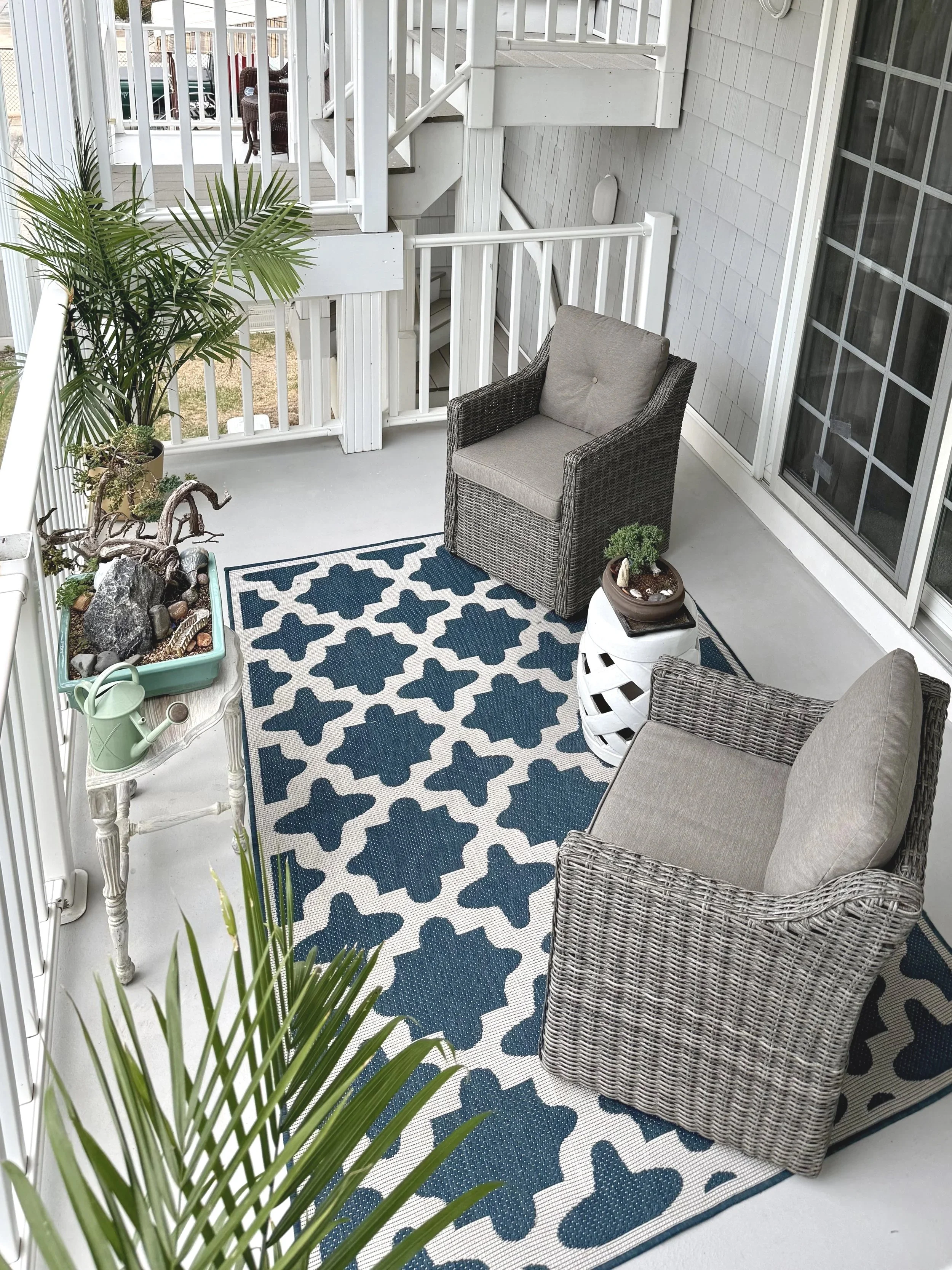 A patio space with wicker and fabric chairs, a patterned blue and white rug, potted plants, and a white railing.