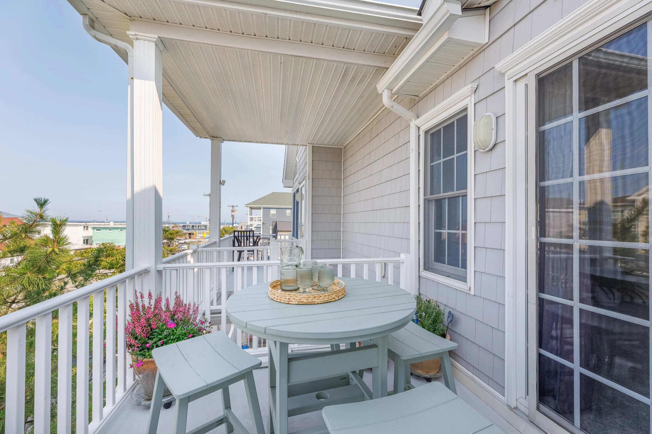 A balcony with a round table, four benches, a tray with glasses, potted flowers, a railing, and neighboring houses under a clear sky.