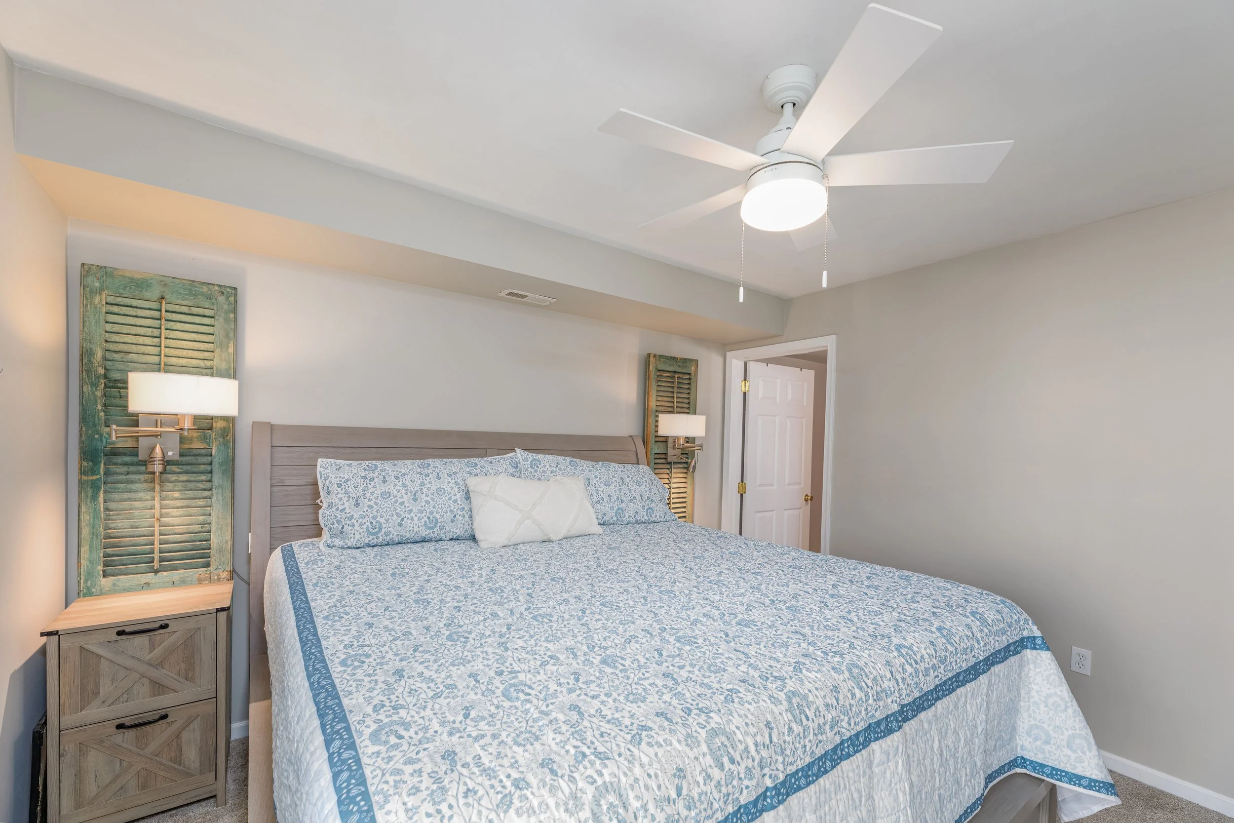 Bedroom featuring a bed with a blue and white patterned quilt, wooden nightstand, and rustic wooden shutters on the wall as headboard. Ceiling fan with light. Light-colored walls.