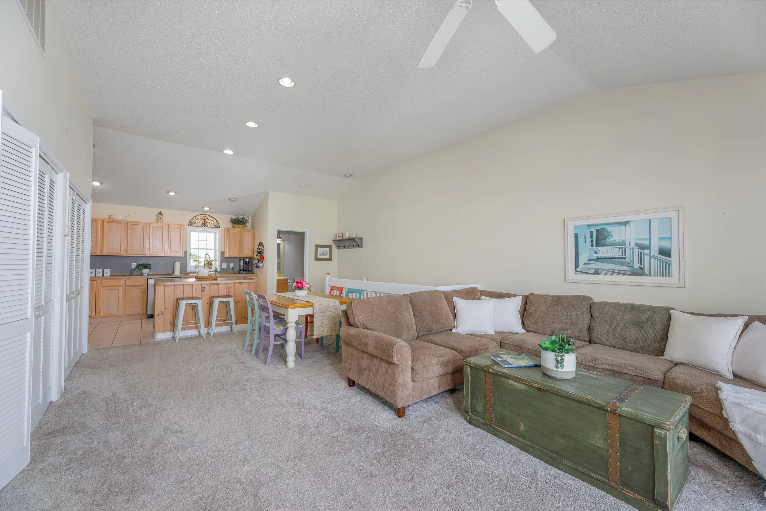 Open-concept living room with beige carpet, brown sectional sofa with white pillows, distressed green coffee table with books, dining area with colorful chairs, wooden kitchen with bar stools, white window, framed nature photo, ceiling fan, and potte