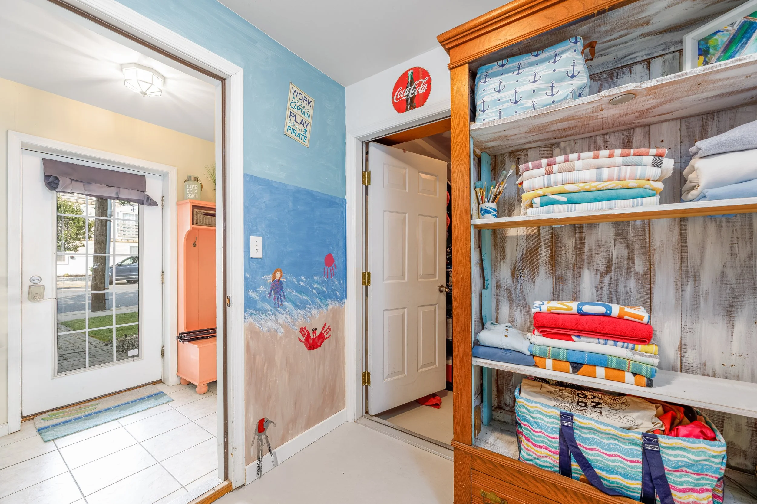 Entryway with glass door leading outside, a wall painted with a blue ocean scene, and a wooden shelving unit filled with neatly folded towels in colorful patterns.