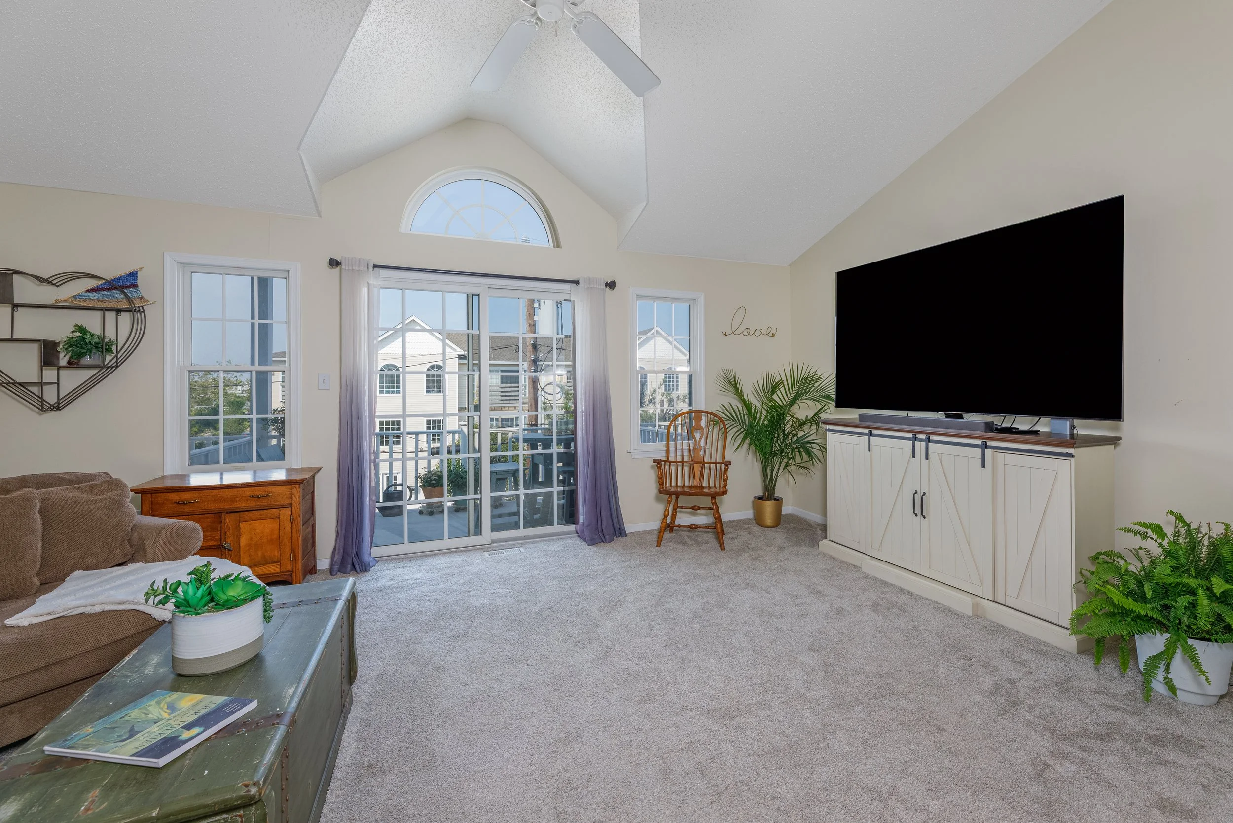 Living room with beige walls, a large flat-screen TV on a white cabinet, a wooden chair, green plants, a sliding glass door with curtains, and a ceiling fan.