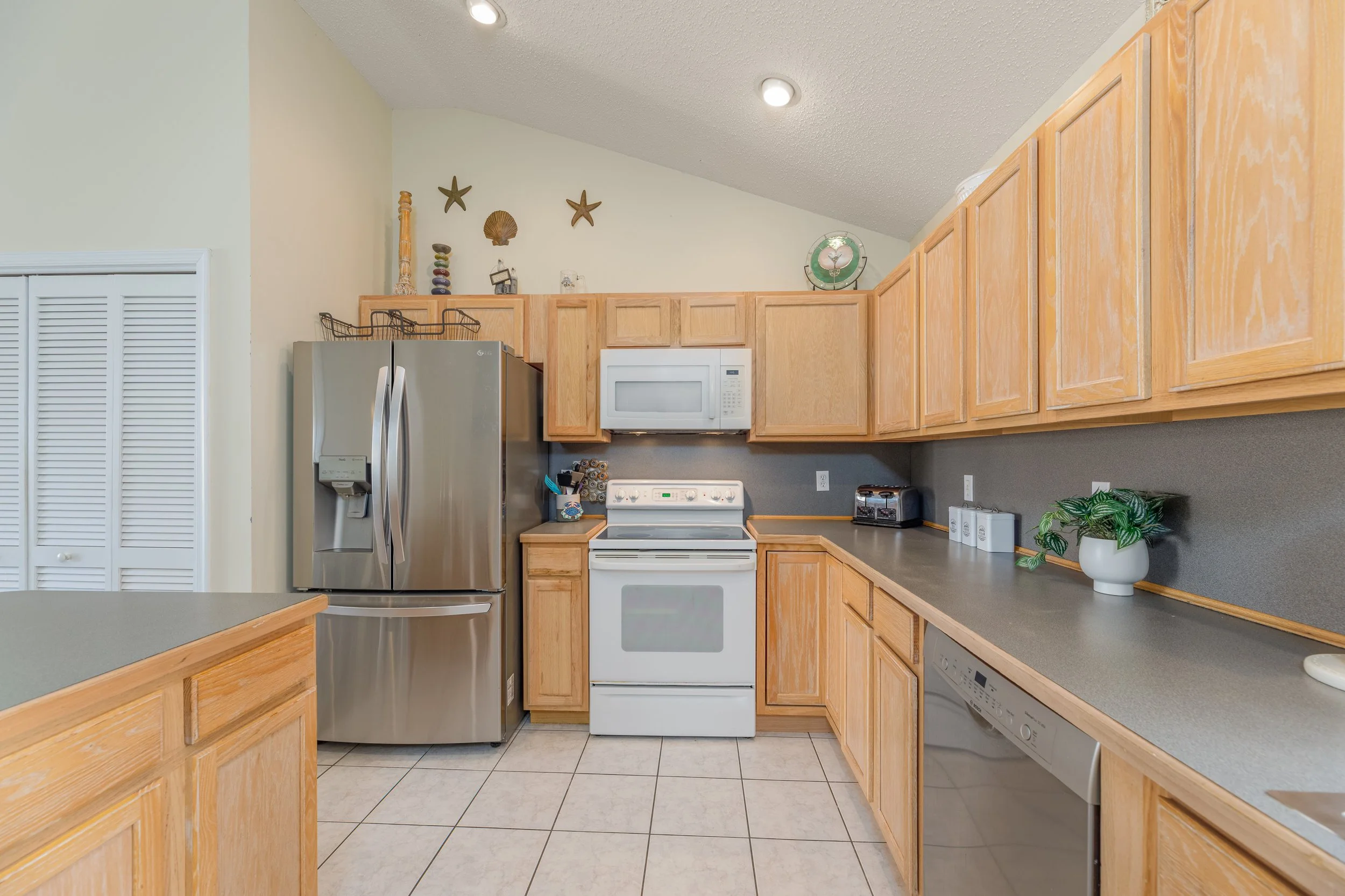 Kitchen with wood cabinets, gray countertops, white appliances including microwave and stove. Stainless steel refrigerator, dishwasher, and a potted plant on the counter, with decorative items and starfish on the wall above the cabinets.