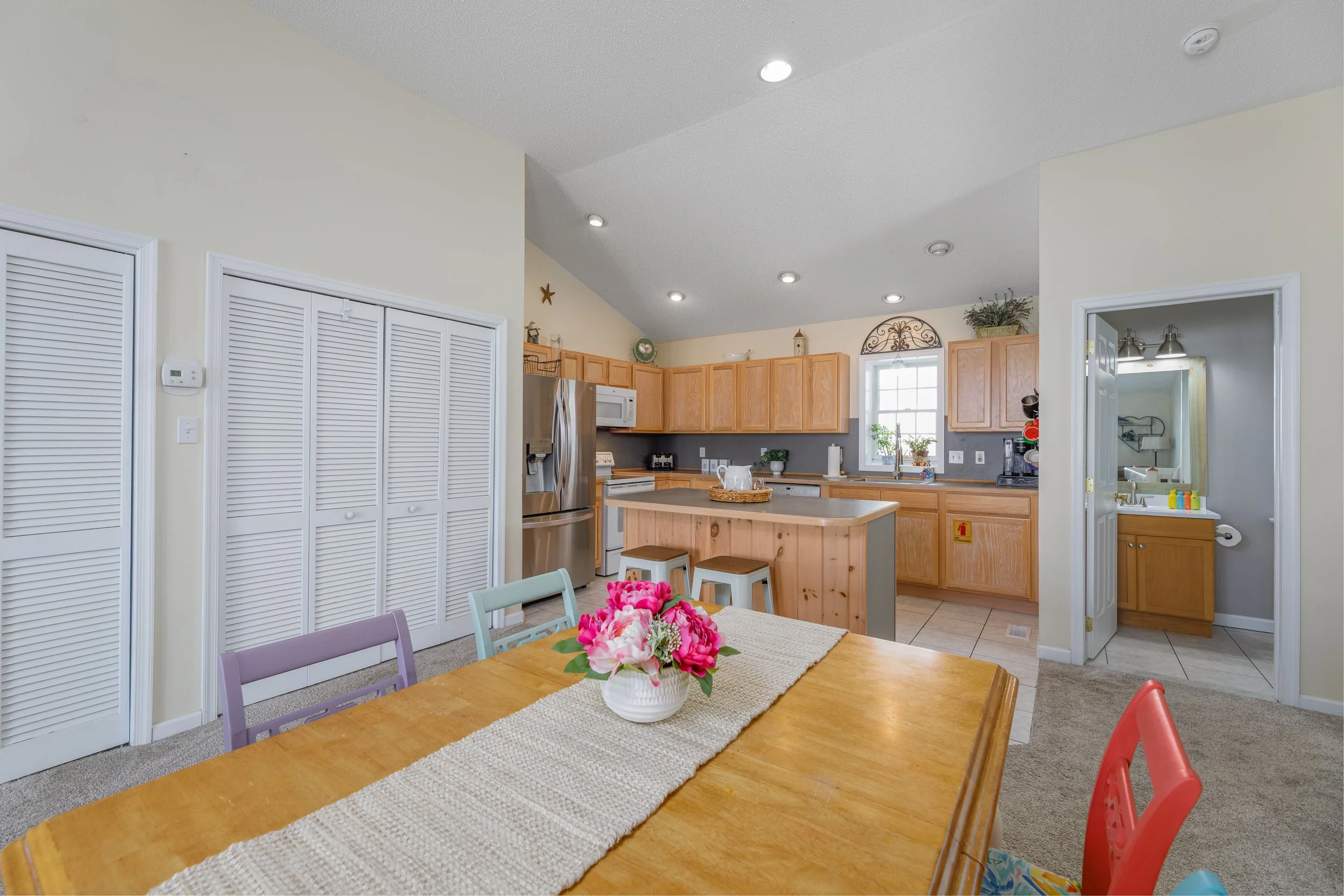View of a kitchen with wooden cabinets, stainless steel refrigerator, microwave, and a dining table with colorful chairs and a flower centerpiece.