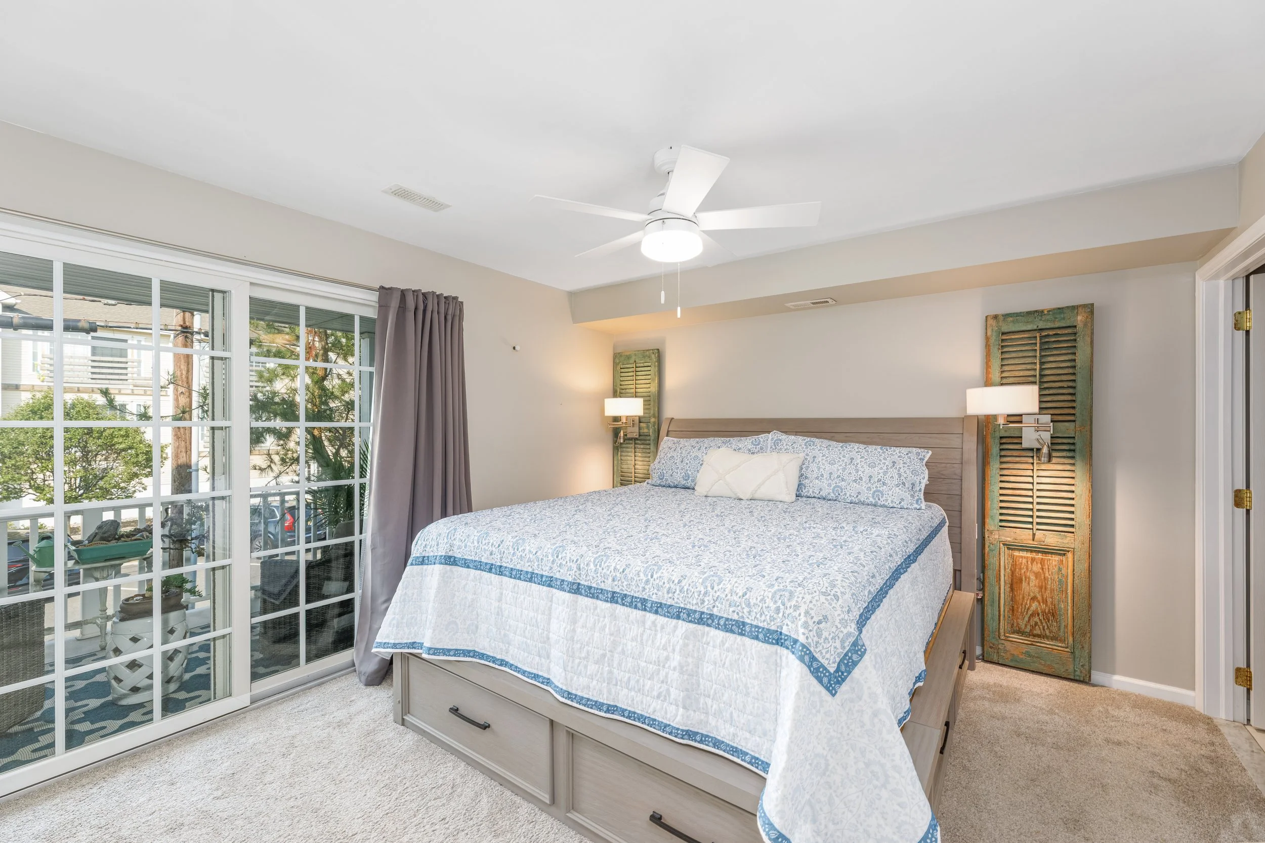 A bedroom with a large bed covered in a blue and white quilt, a wooden headboard, and a decorative green and brown shutter on the wall behind it. A sliding glass door with a grid pattern leads to a balcony with outdoor furniture and plants. The room 