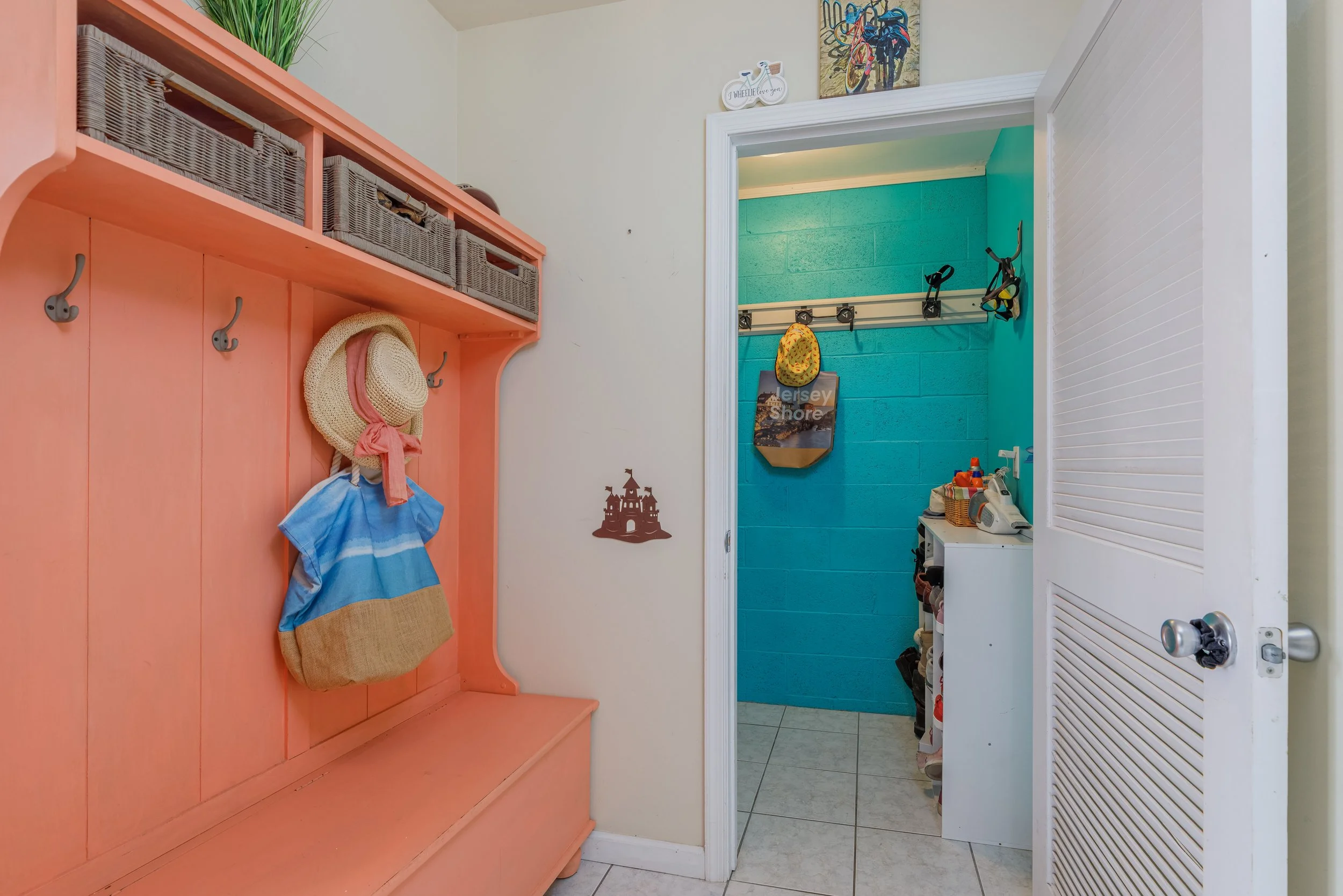 A glimpse into a mudroom or entryway features a pink wooden bench with coat hooks and a woven basket hat. The wall above has a shelf with wicker baskets and decorative items. A doorway leads to a small laundry or utility room with bright teal walls, 