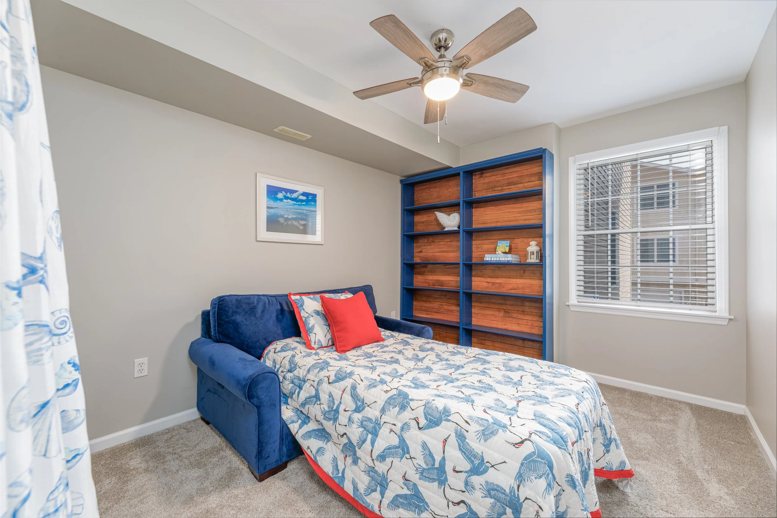 A bedroom with a bed covered in a blue and white patterned bedspread featuring sailboats, a red pillow, and a floral pillow. There is a blue and brown wooden bookshelf against the wall, with decorative items on it. A window with white blinds is on th