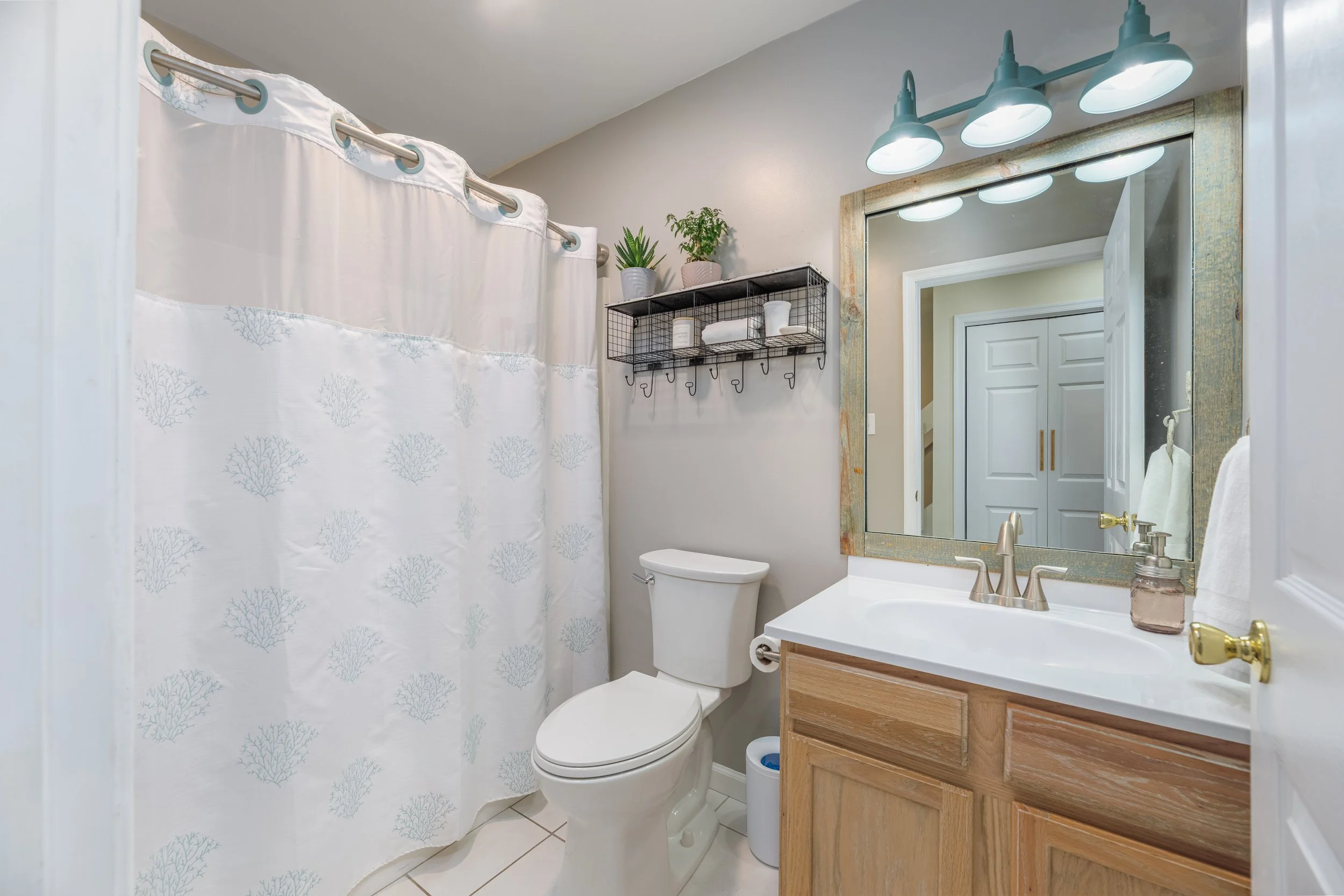 Small bathroom with a white toilet, a wooden vanity with a white countertop, a mirror with a three-light fixture above, a towel ring with a towel, a black metal shelf with plant pots and towels, and a shower curtain with a subtle coral pattern.