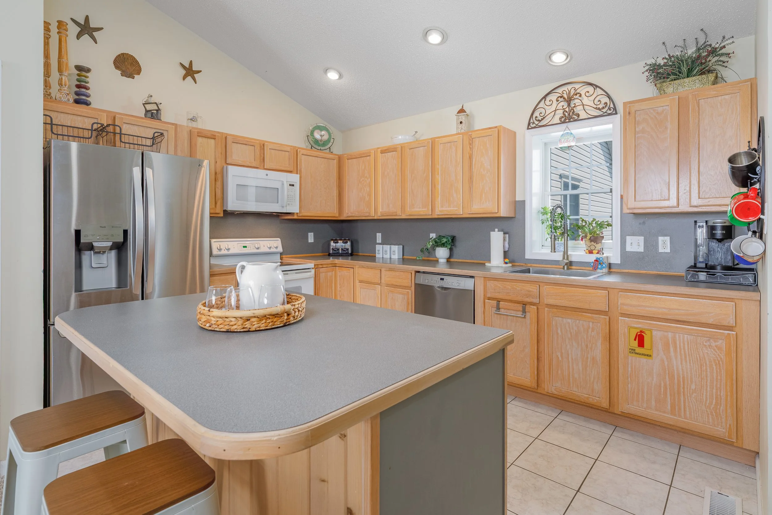 Kitchen with wooden cabinets, stainless steel refrigerator, microwave, dishwasher, and a kitchen island. Decor includes wall plates and starfish, potted plants, and hanging mugs by the window.
