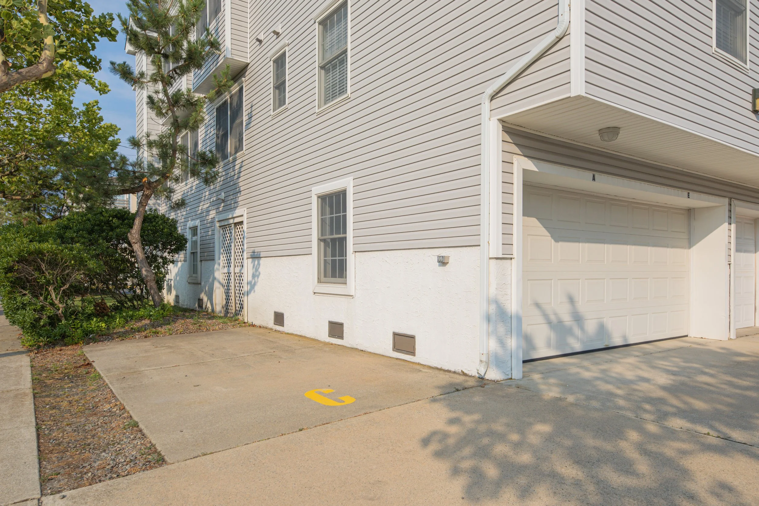 A beige residential building with white trim and multiple windows, a garage door, and an attached carport. There is a tree with green foliage and bushes in front, and the quoted letter C is painted on a parking space in the concrete driveway.