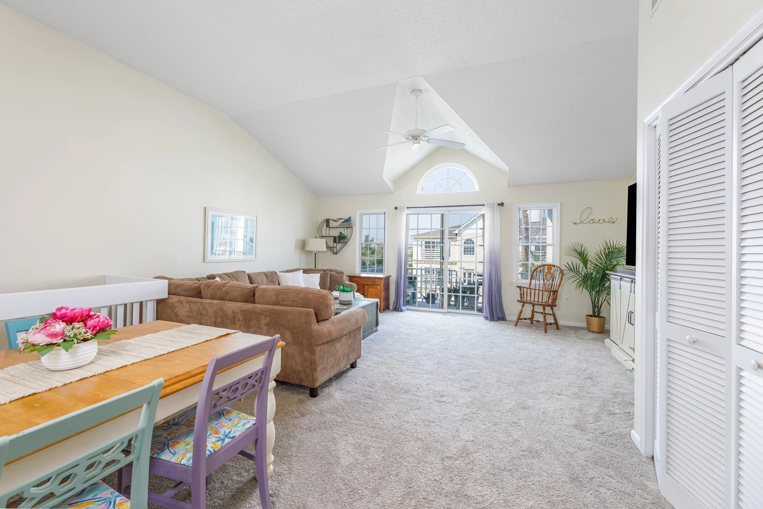 Living room with beige carpet, beige sofa, wooden coffee table, white walls, large windows with sheer purple curtains, sliding glass door, ceiling fan, and white louvered closet doors.