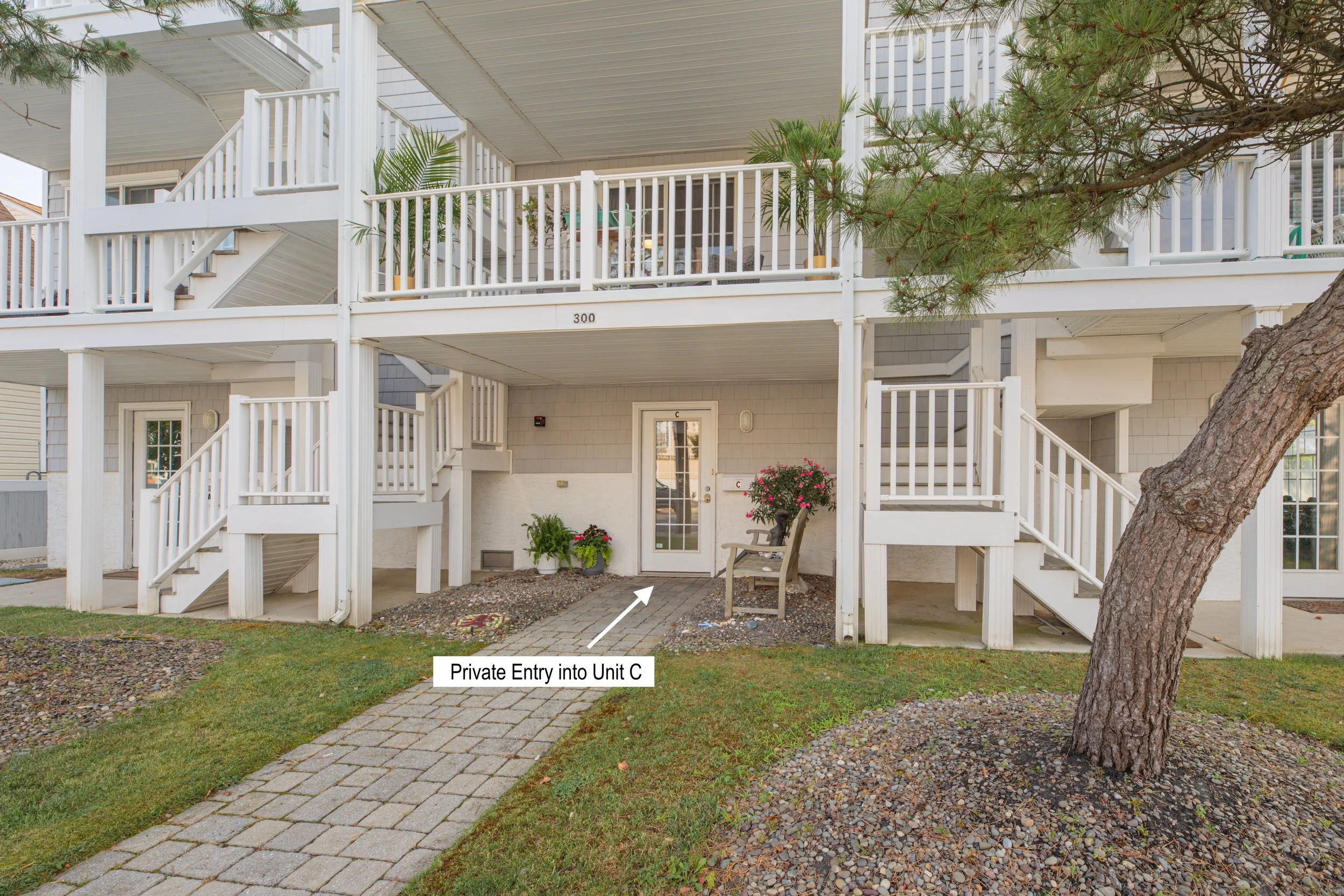 Exterior view of a multi-level residential building with white railings and stairs, a large tree, a bench with pink flowers, and a concrete pathway. There is a label indicating a "Private Entry into Unit C."