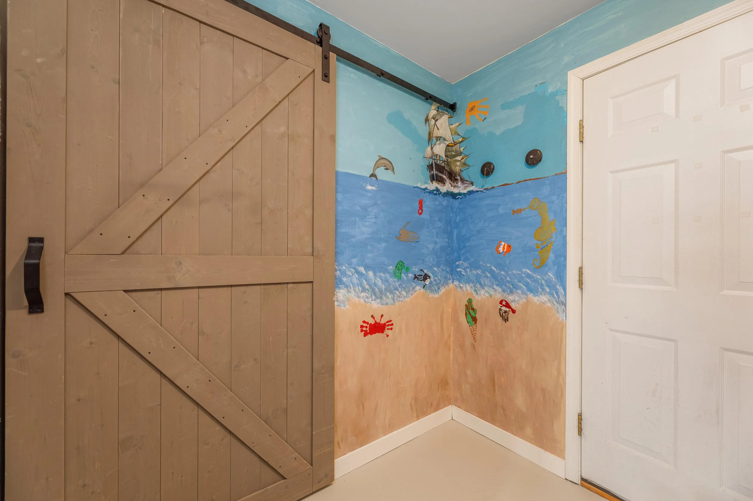 A corner of a room with a wooden sliding barn door on a black metal track. The wall has a painted mural of an ocean scene with a ship, dolphins, fish, a seahorse, and crabs.