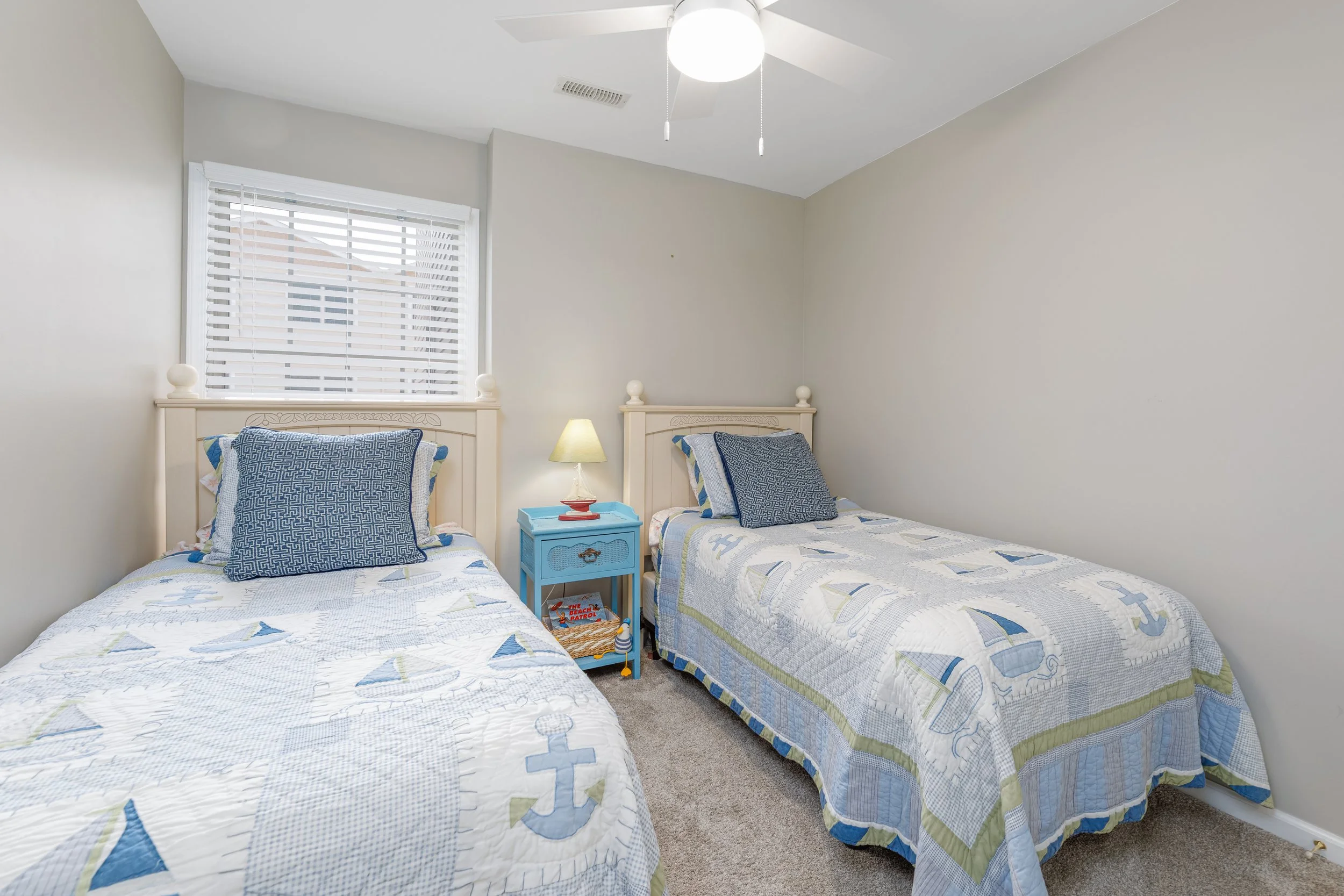 Two twin beds with matching nautical-themed bedspreads and pillows in a bedroom with a window, a blue nightstand, a small lamp, and a ceiling fan.