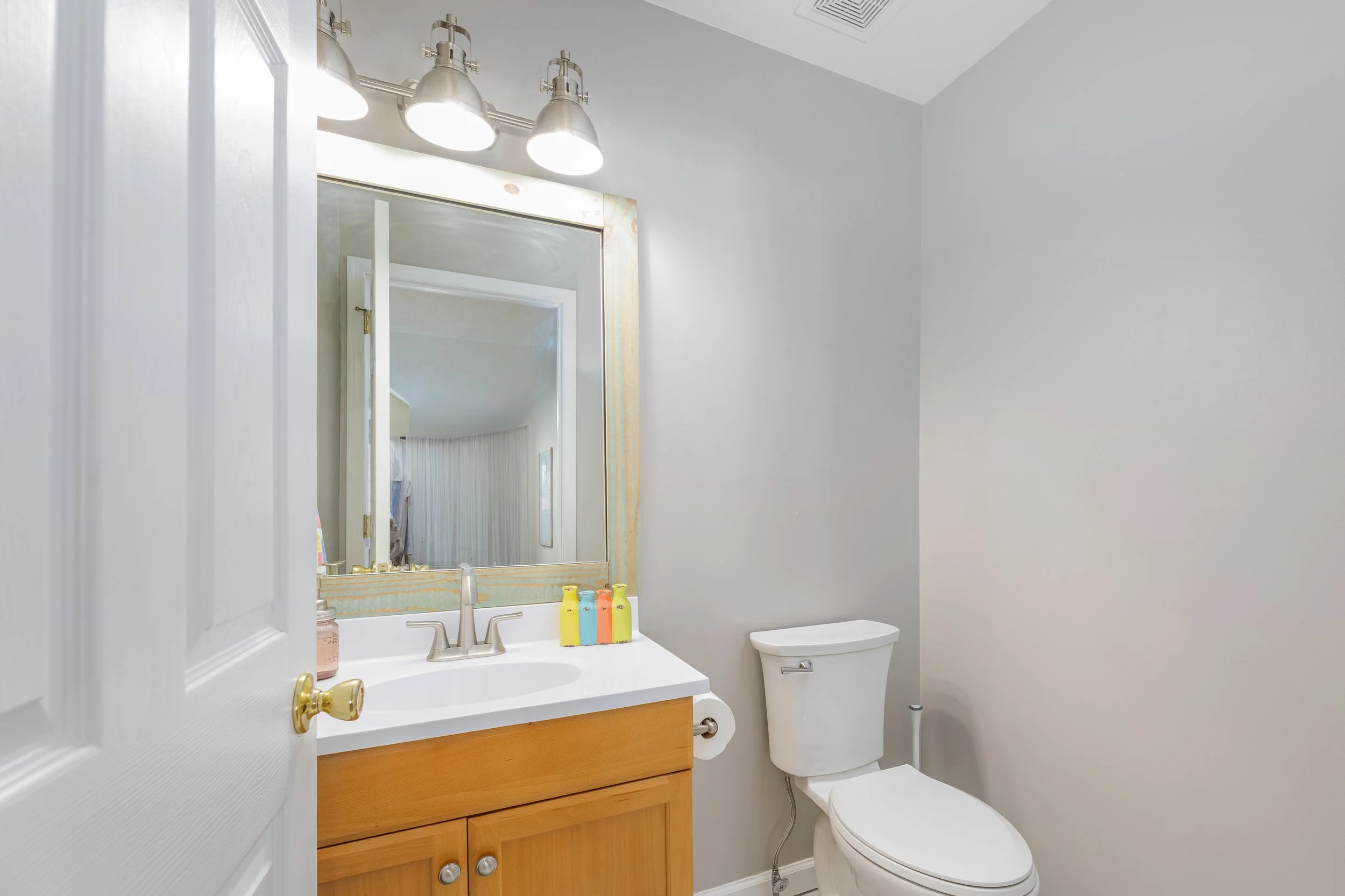 A bathroom with a white sink, wooden cabinet, mirror, light fixture with three bulbs, toilet, and pastel-colored bottles on the sink
