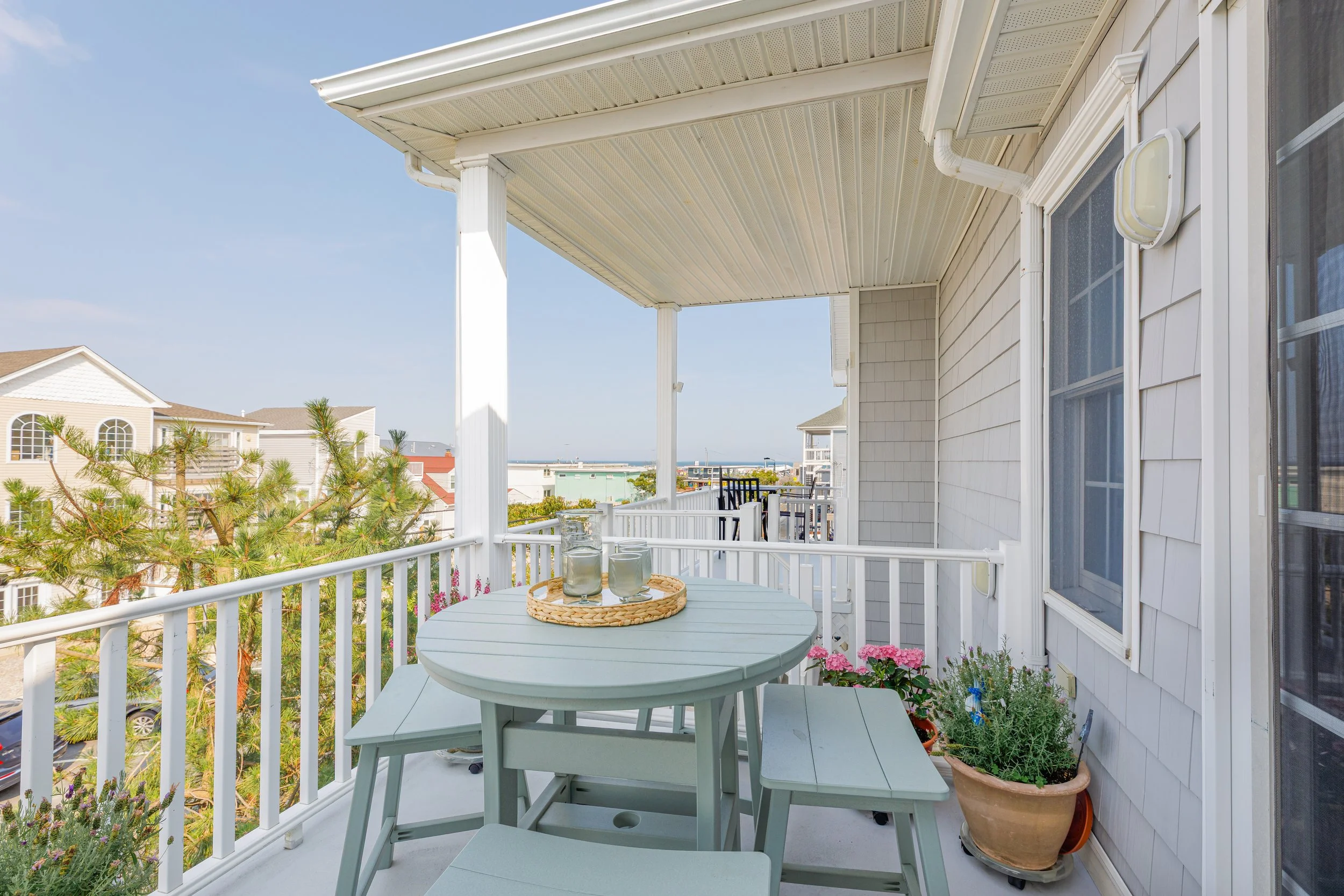 Balcony with white railing, patio table with candles, potted plants, and outdoor view of neighboring houses and ocean under a clear blue sky.
