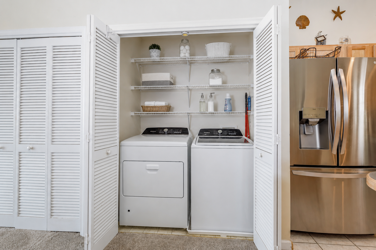 A laundry area with a white washing machine and dryer behind white louvered closet doors, and shelves with laundry supplies and decorative items above the appliances. To the right is a stainless steel refrigerator.