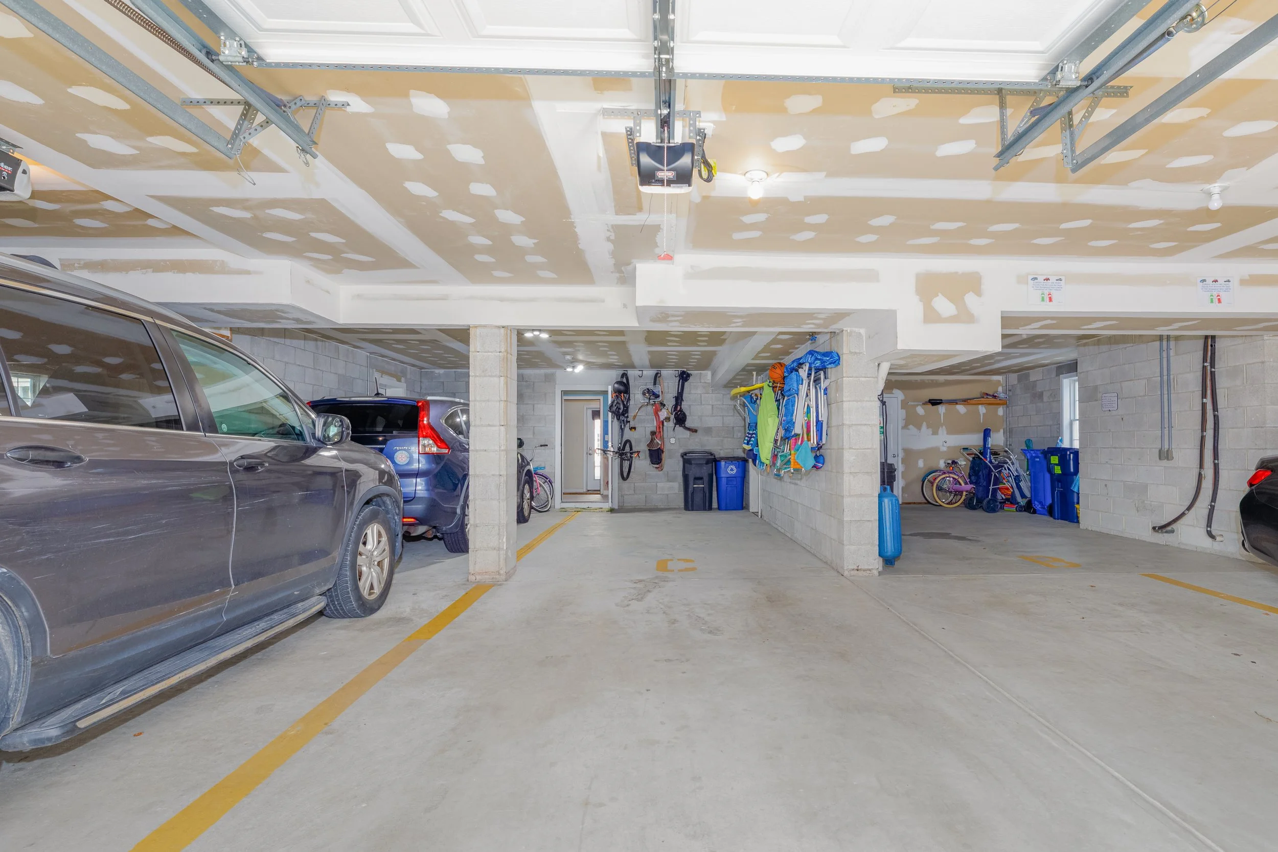 Underground parking garage with parked cars on the left, unfinished drywall on the ceiling, and storage items such as bicycles, bins, and equipment on the right.