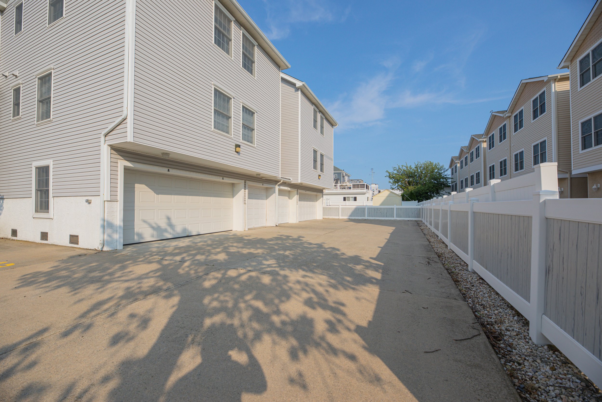 Row of beige townhouses with attached garages, white fences along the sidewalks, and a parking area in front, under a clear blue sky.