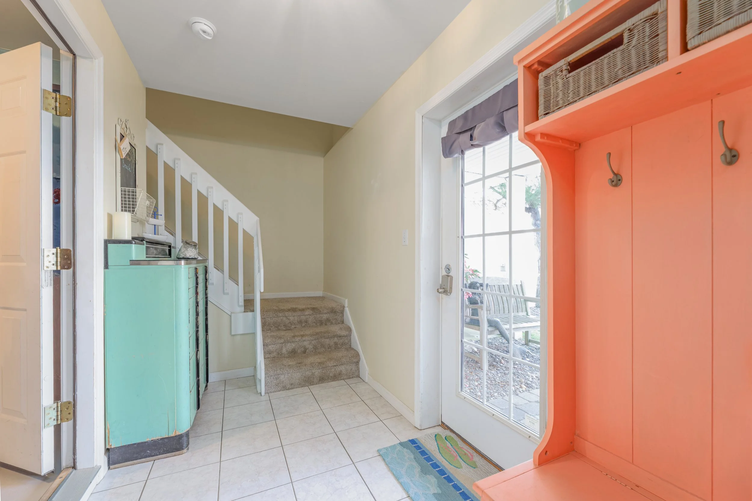 Entryway with tiled floor, colored cabinets, and a sliding glass door leading outside where patio furniture is visible. A small staircase with carpeted steps, a partially open door, and a light-colored wall are also visible.