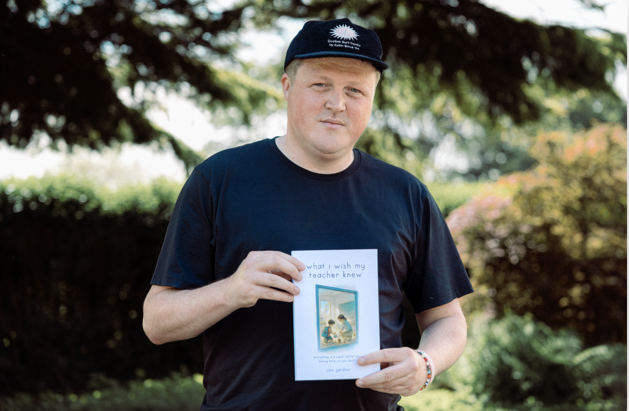 A young man with a serious expression wearing a black t-shirt, a black hat with white text, and a colorful bracelet on his right wrist. He is holding a book titled "what I wish my teacher knew" outdoors with trees and greenery in the background.