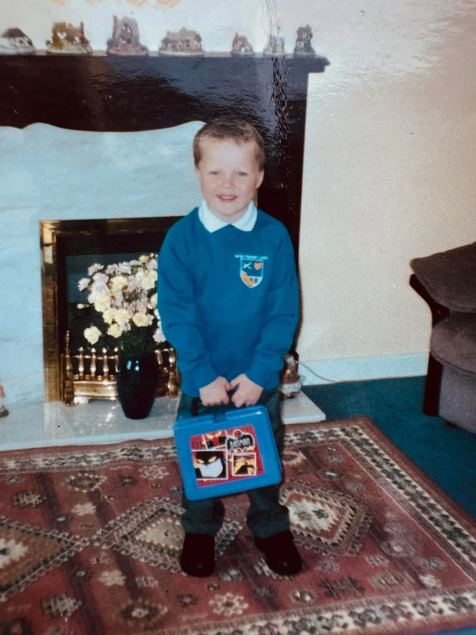 A young boy with short light brown hair, wearing a blue school sweater with a crest on it, stands in a living room. Behind him, a framed picture of flowers, a tall black vase, a small decorative jar, and a Pop Art cardboard case with comic strip images are on a white table. The room has a patterned rug on the floor, a beige wall, and a black chair on the right side.