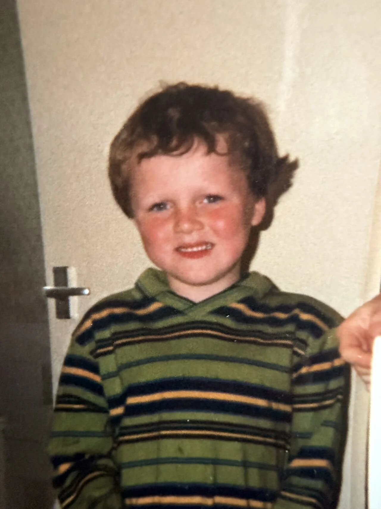 A young boy with short, curly hair and blue eyes is smiling slightly while wearing a green and black striped hoodie. He is standing indoors near a beige wall and a door.