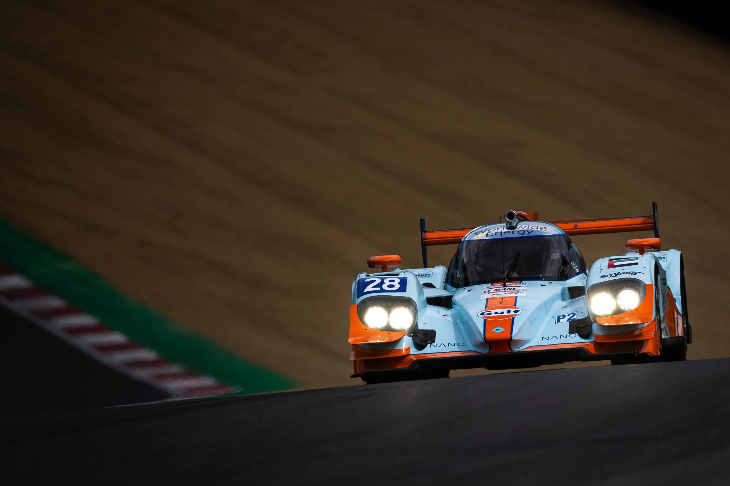 A racing car with the number 28 on it navigating a turn on a racing track at night. The car features Gulf Oil sponsorship and has bright headlights illuminating the track.