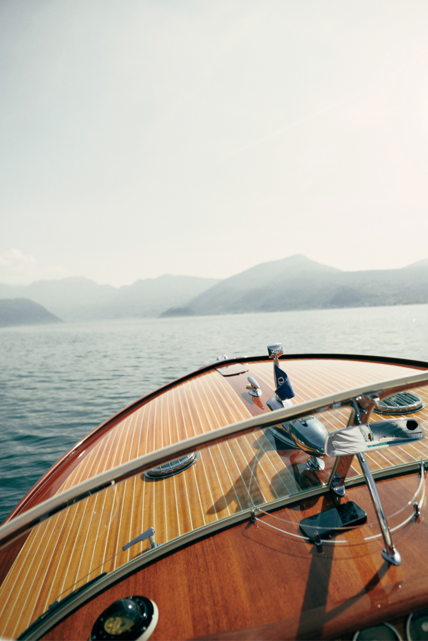 View from the front of a boat with a wooden deck, showing the boat's windshield, hardware, and a calm body of water with distant mountains under a clear sky.