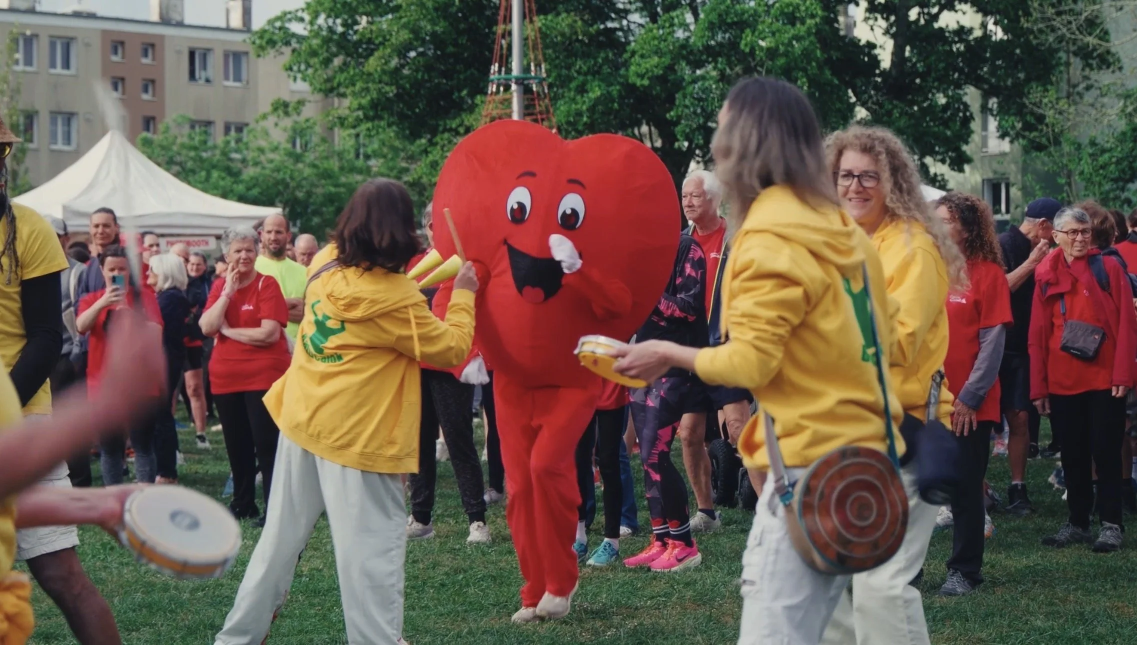 Fête en plein air avec un personnage en costume de cœur rouge souriant entouré de personnes portant des vêtements jaunes, dans un parc avec des arbres et des bâtiments en arrière-plan.
