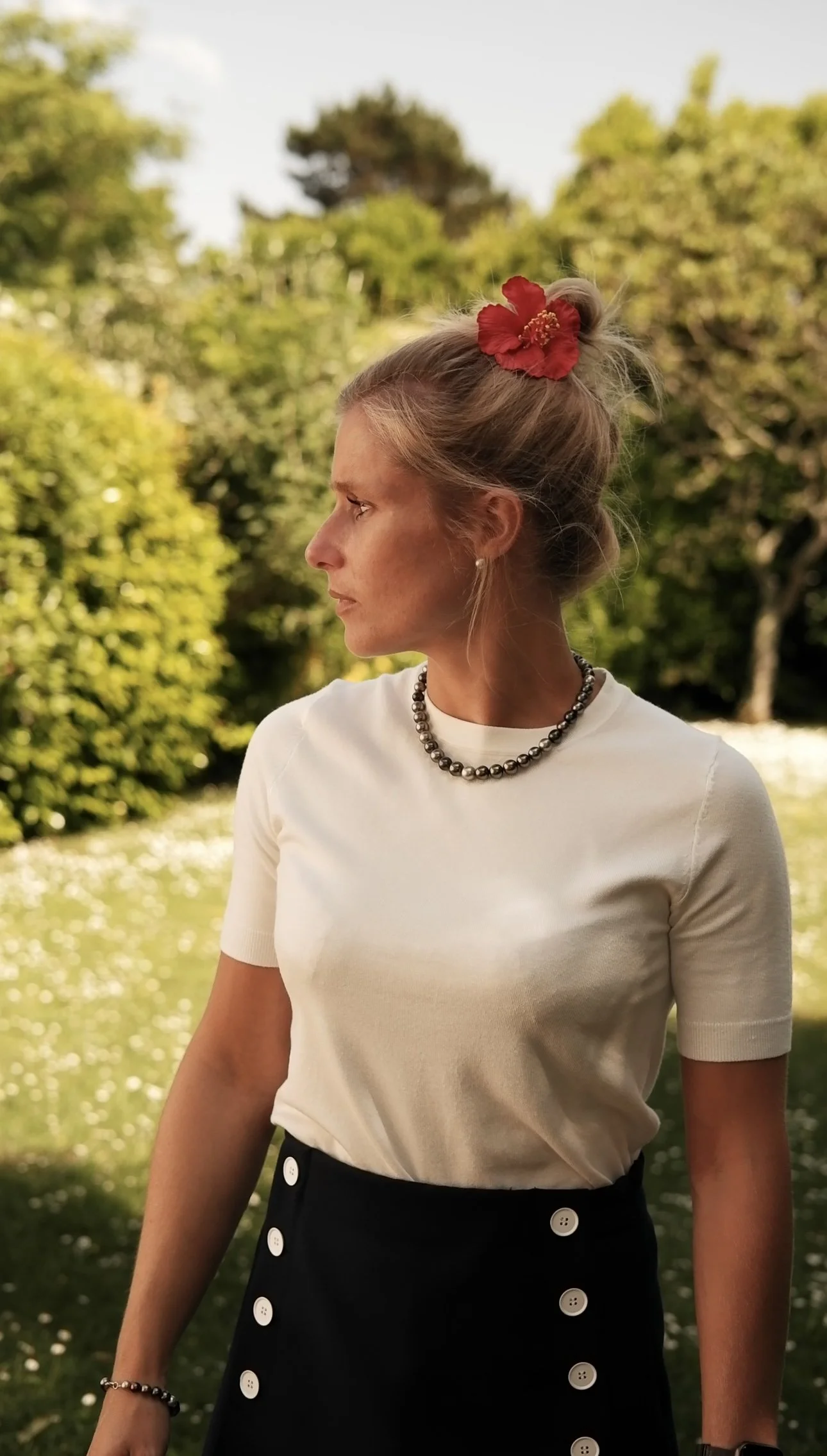 Femme blonde portant un haut blanc, une jupe noire avec boutons blancs, un collier de perles, une montre et un bracelet, avec une fleur rouge dans ses cheveux, dans un parc en extérieur