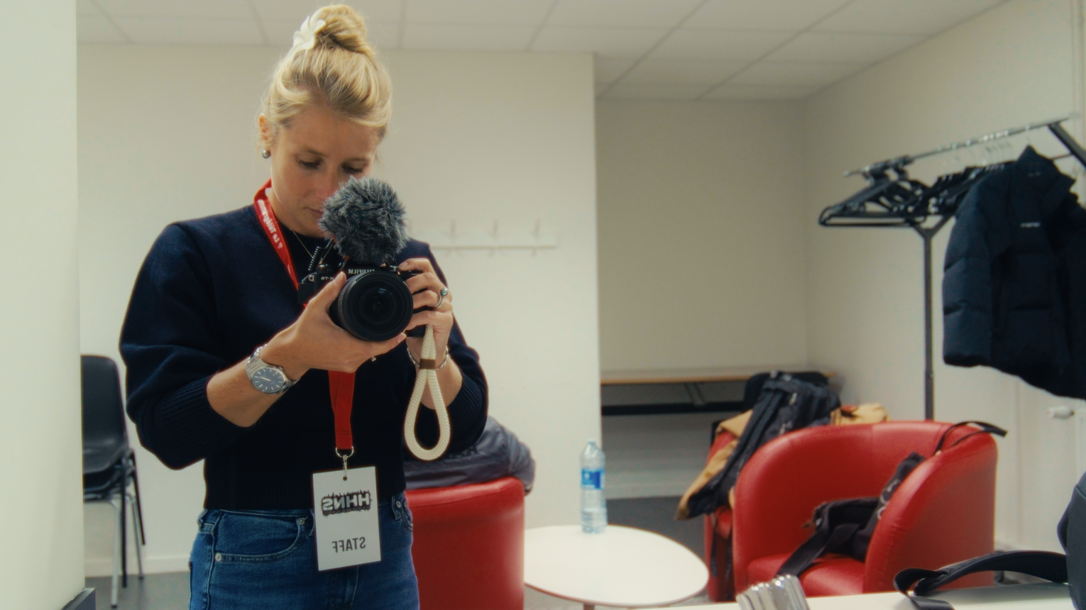 Femme prenant une vidéo avec une caméra dans une salle de réunion ou de pause, avec des fauteuils rouges, une table, un sac à dos, une bouteille d'eau, des vêtements suspendus et un mur blanc.