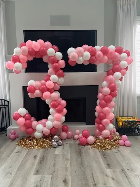 Large pink and white balloon arrangement shaped into the number 30, decorated with gold confetti at the base, in front of a fireplace and television in a living room.