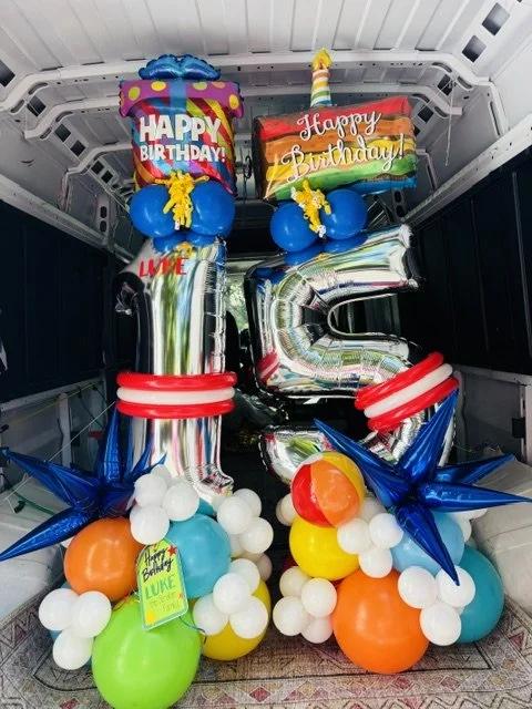 Balloon arrangement celebrating a first birthday, including large number 1 balloons, a cake shaped foil balloon with birthday messages, and clusters of colorful smaller balloons.