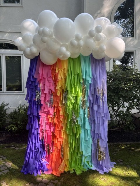 Decorative rainbow with multicolored streamers and white balloons arranged to resemble clouds is a colorful party balloon backdrop for a 7th birthday party in Newtown PA.