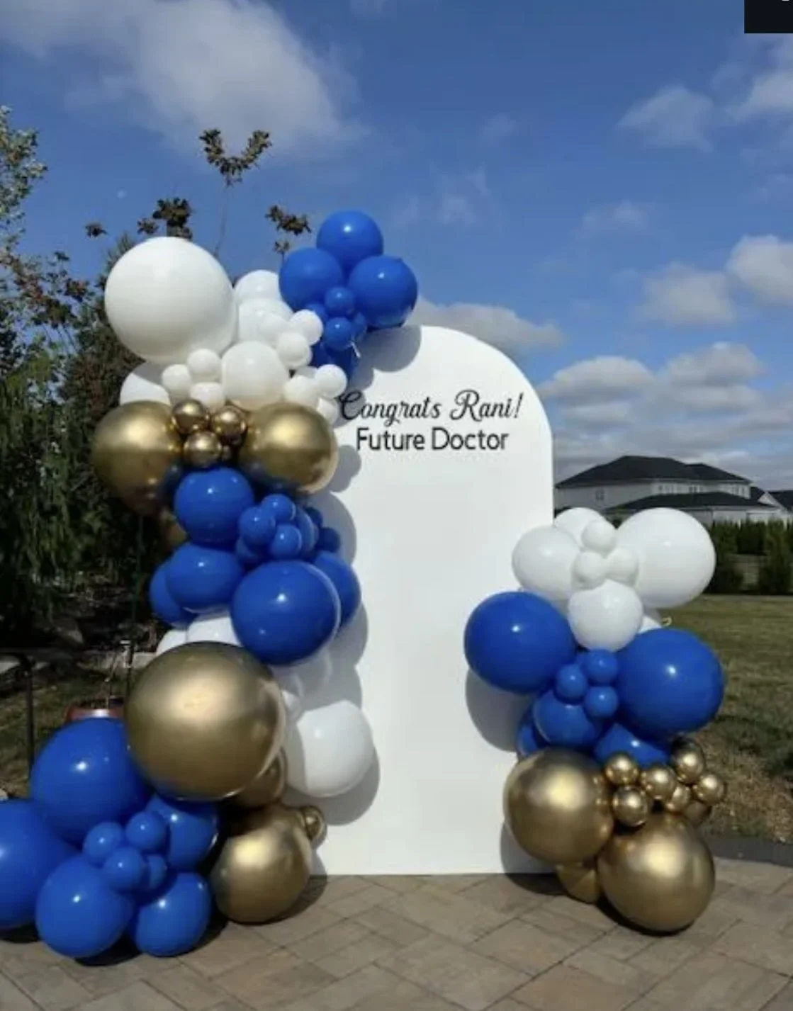 A decorative outdoor display with a white board surrounded by clusters of blue, white, and gold balloons. The board reads "Congrats Rani! Future Doctor" against a background of a blue sky with clouds, trees, and a house.