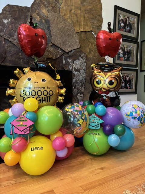 A colorful balloon display including an owl-shaped balloon, apple-shaped balloons, a globe, and a yellow balloon with 'someday future is SOOOO BRIGHT!' written on it, set against a stone wall with framed photos, and featuring pink alert tags with the