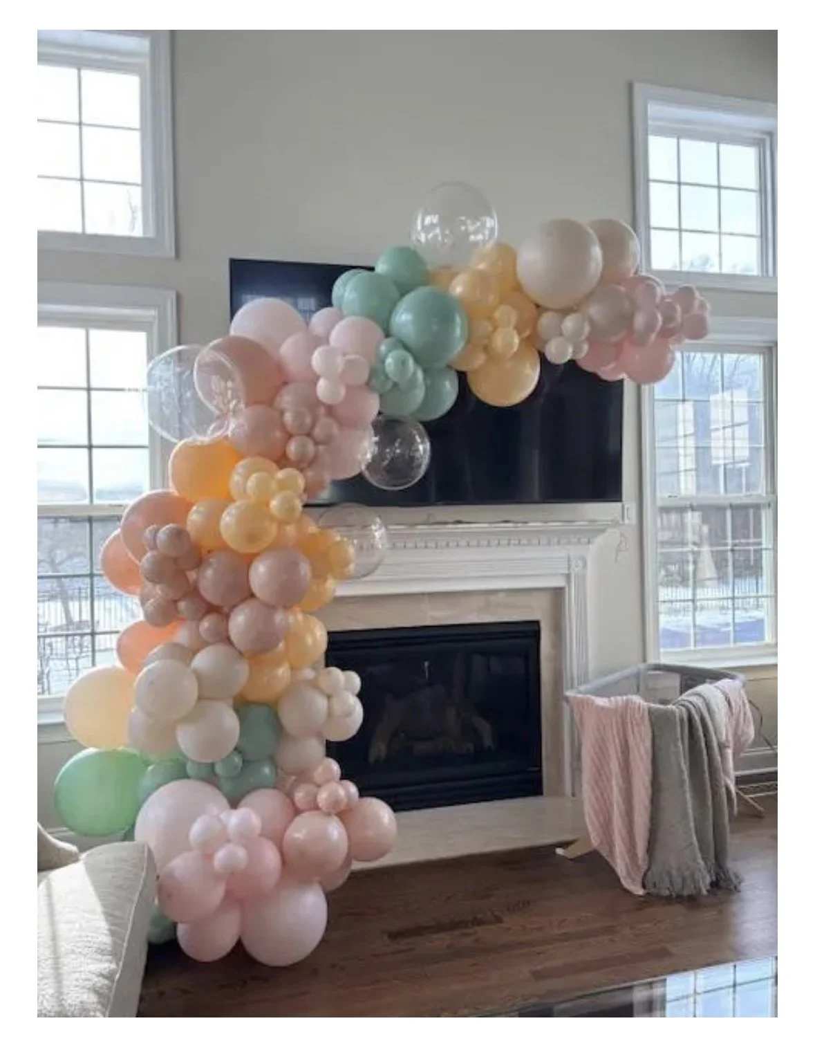 Colorful balloon arch with pink, peach, green, and clear balloons decorating a living room with large windows and a fireplace.