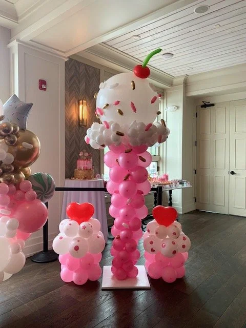 Balloon sculpture of ice cream sundae with a cherry on top, surrounded by balloon roses with red heart toppers, in a decorated indoor setting.
