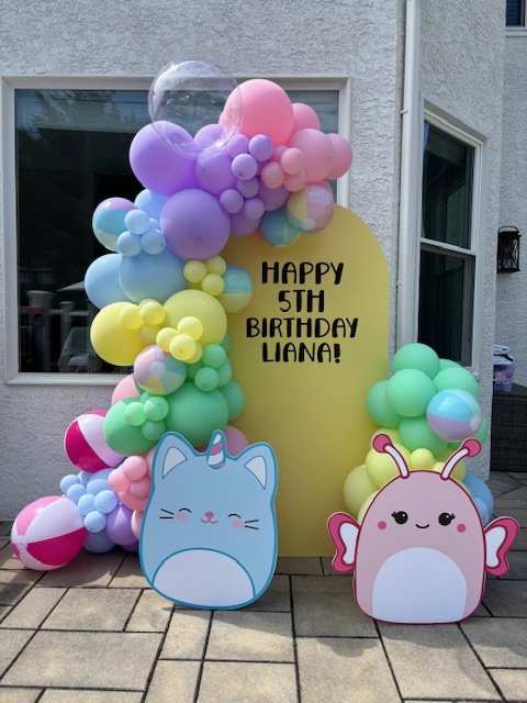 Colorful balloon arch with pastel pink, purple, yellow, blue, and green balloons, featuring a yellow sign that says "Happy 5th Birthday Lana!" in black letters. Two large cartoon cutouts of a unicorn and a butterfly with smiling faces are in front of