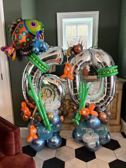 Balloon sculpture with large silver foil numbers '20', decorated with colorful animal and sea creature balloons, set in front of a window in a room with green walls and a hexagonal tile floor.